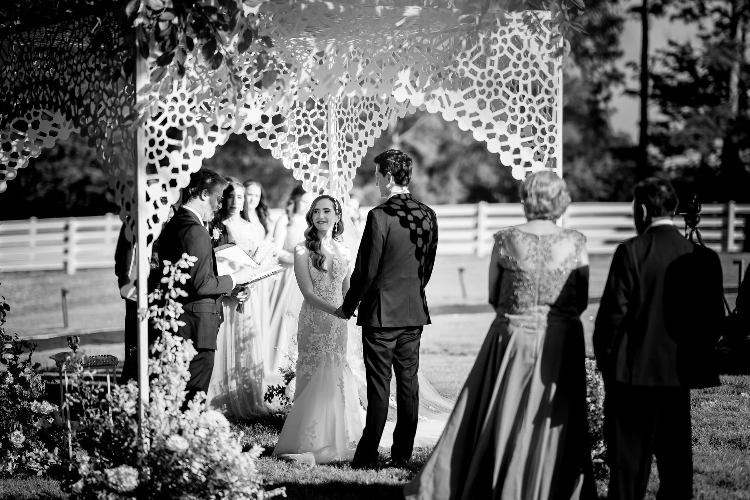 historic wakefield barn wedding photography - jennifer & stefan A bride and groom stand under a decorated gazebo during a charming Wakefield barn wedding ceremony. The bride, in her white gown, holds hands with the groom. Bridesmaids and guests look on, as stunning floral arrangements surround the scene, vividly captured in wedding photography.