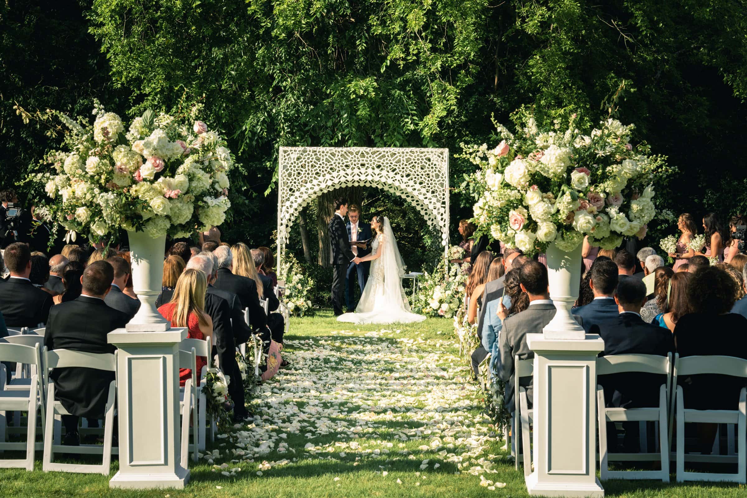 A bride and groom stand under a white floral arch during an outdoor wedding ceremony at Wakefield. Guests sit on either side, facing the couple. The aisle is adorned with scattered rose petals and large floral arrangements. Lush green trees are visible in the background, perfect for barn wedding photography.