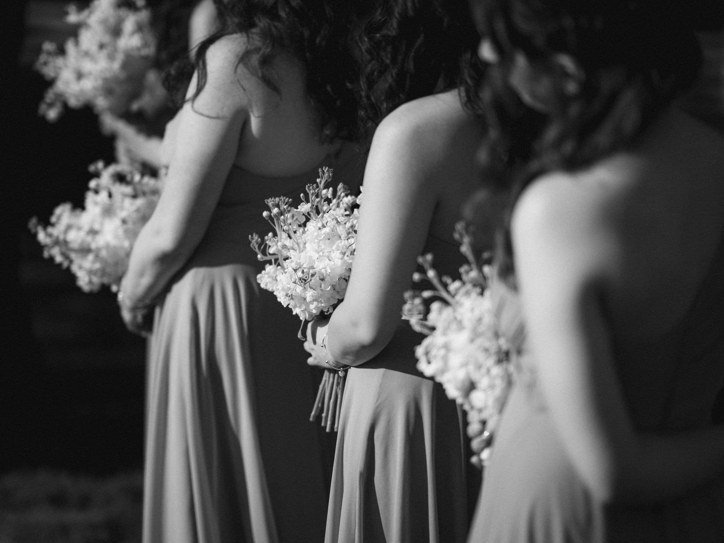 historic wakefield barn wedding photography - jennifer & stefan Four women in elegant dresses stand in a line at a Wakefield barn wedding, each holding a bouquet of flowers. The black and white image beautifully highlights the flowing fabric and delicate floral arrangements, capturing the timeless charm of barn wedding photography.