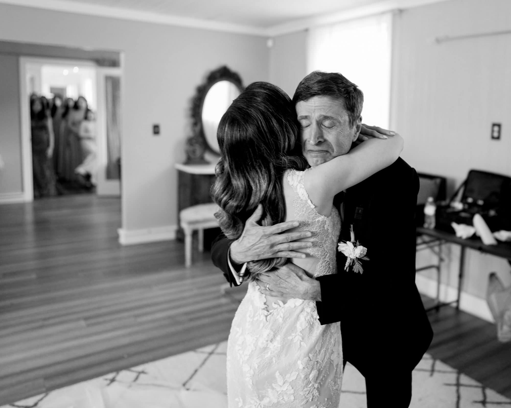 historic wakefield barn wedding photography - jennifer & stefan In an emotional and intimate scene at Wakefield Barn, a bride in a lace wedding dress hugs an older man in a suit inside a room with elegant furniture. A group of people in formal attire watches from the doorway, capturing the essence of barn wedding photography.