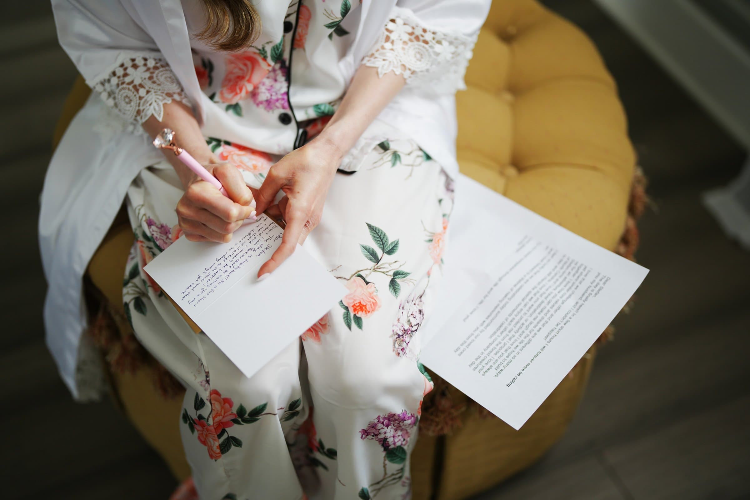 historic wakefield barn wedding photography - jennifer & stefan A person in floral pajamas sits on a yellow cushion, writing on a card with a pink pen. Nearby are two sheets of paper, one partially visible. The scene feels reminiscent of tranquil moments captured in Wakefield barn wedding photography.