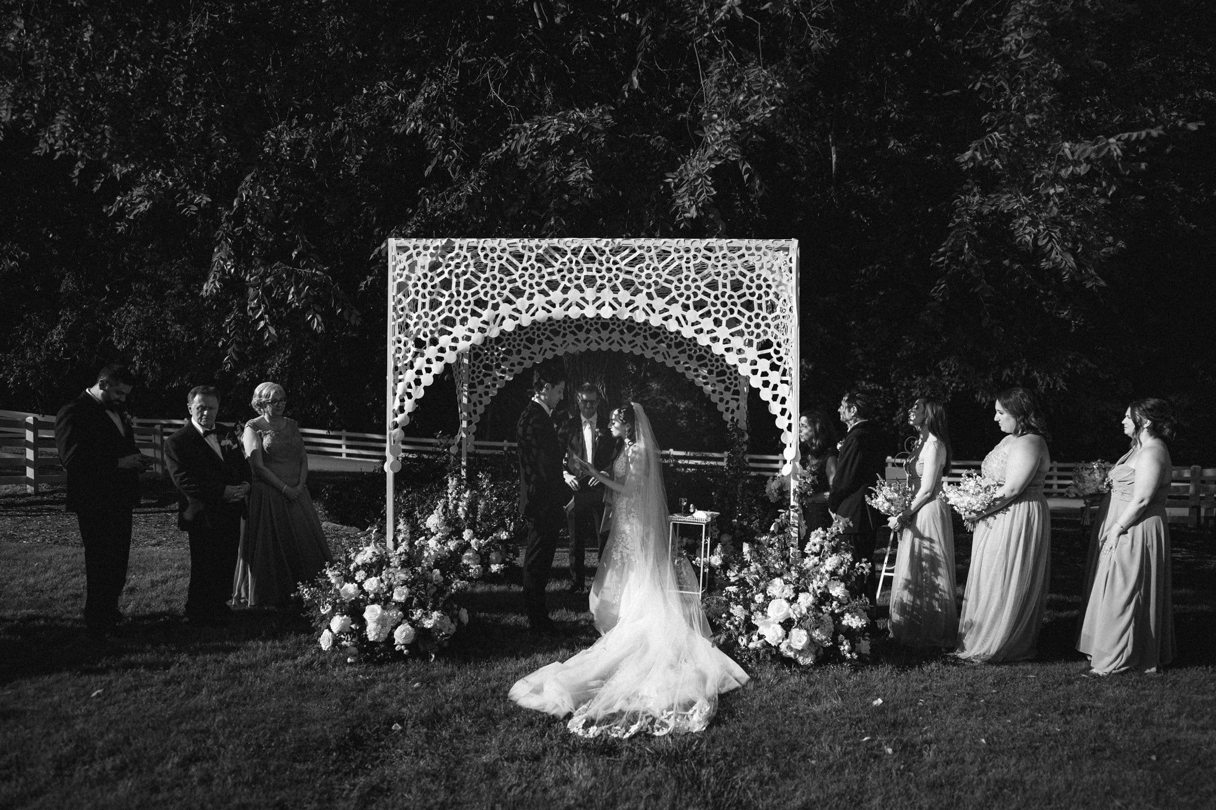 historic wakefield barn wedding photography - jennifer & stefan A black and white photo captures a Wakefield barn wedding ceremony. The couple stands elegantly under a decorative floral arch, flanked by attendants. Guests are seated on the grass, with trees as a serene backdrop. The bride's long veil and gown complete this timeless scene.