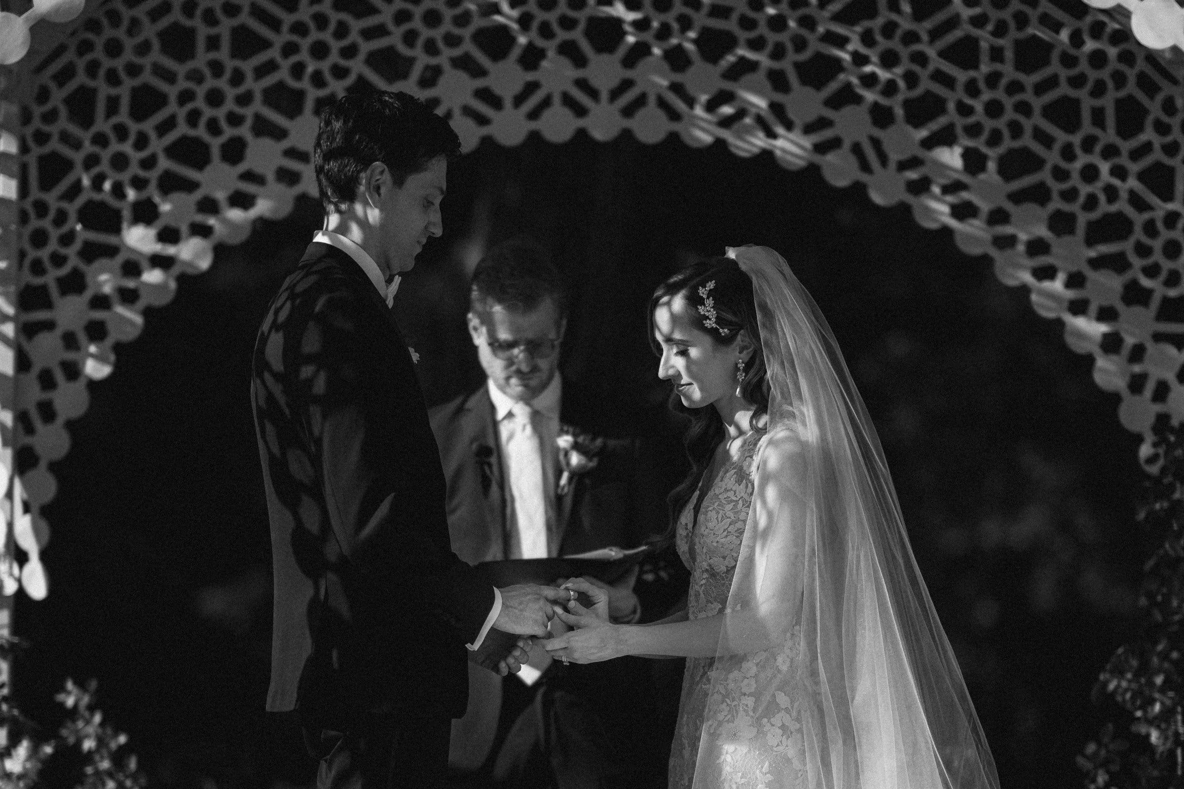 A black and white photo captures a Wakefield barn wedding ceremony. The couple stands facing each other, holding hands under a decorative arch. The groom wears a suit, and the bride is in a lace gown with a veil, as the officiant stands behind them holding a book.