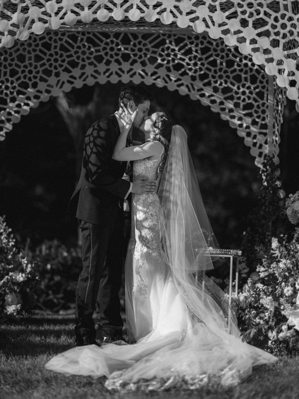 historic wakefield barn wedding photography - jennifer & stefan A newlywed couple shares a kiss under a decorative arch at an enchanting Wakefield barn wedding. The bride, in a long flowing gown and veil, stands beside her suited groom amid flowers and greenery, capturing a timeless moment of romance.