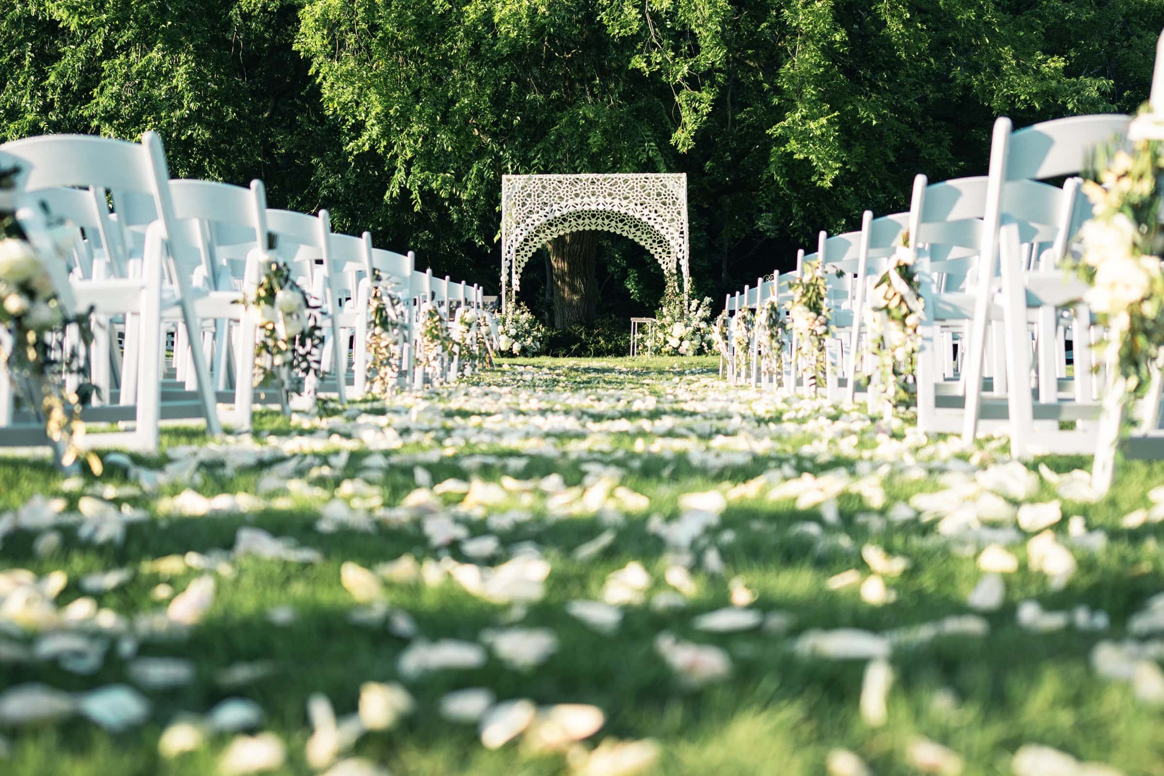 historic wakefield barn wedding photography - jennifer & stefan A picturesque outdoor wedding ceremony setup with white chairs lined up on either side of an aisle adorned with flower petals. Completing the scene is a decorative white arch framed by lush green trees, capturing the essence of Wakefield barn wedding photography.