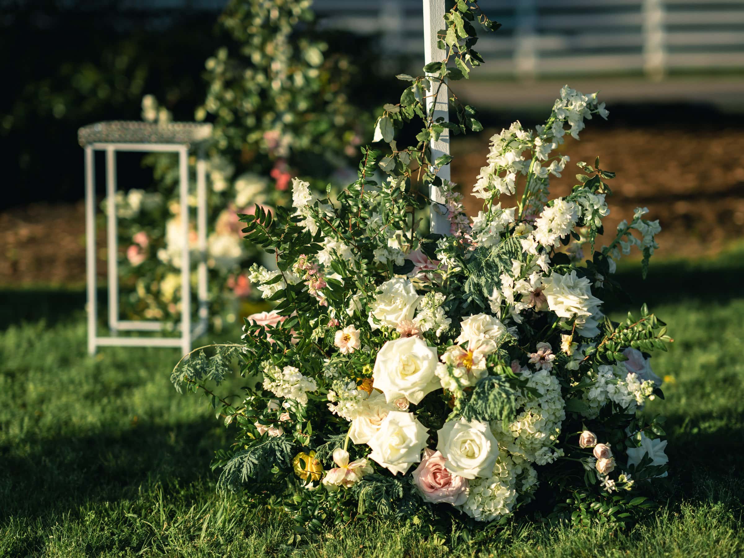 historic wakefield barn wedding photography - jennifer & stefan A lush garden display featuring an arrangement of white and pink roses, surrounded by greenery, is set on a grassy lawn under the soft evening light, perfect for Wakefield barn wedding photography. A white metal lantern stands gracefully in the background, completing the enchanting scene.