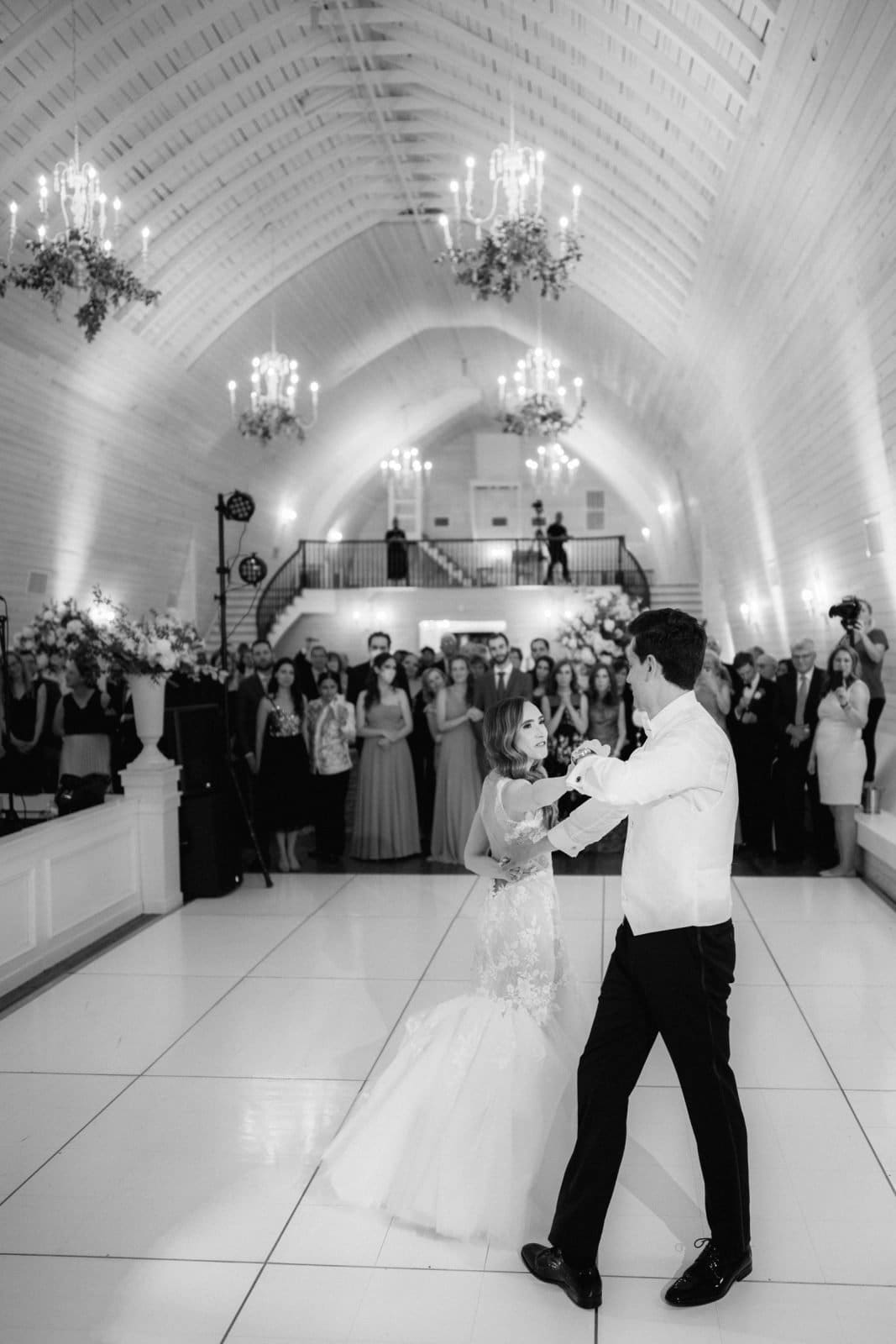 historic wakefield barn wedding photography - jennifer & stefan In the elegantly decorated hall of the Wakefield barn, chandeliers and floral arrangements create a stunning backdrop as a couple dances gracefully. The bride glows in her white gown, while the groom looks sharp in his suit. Guests watch from the sides and a balcony, capturing every moment like perfect wedding photography.