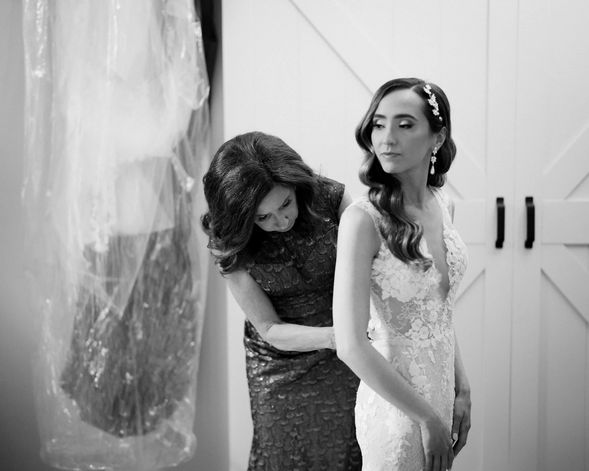 historic wakefield barn wedding photography - jennifer & stefan A black and white photo captures the elegance of a Wakefield barn wedding, with a bride in a lace gown getting ready. Assisted by another woman, she looks forward as a dress hangs nearby in a plastic cover, evoking the timeless charm of this special day.