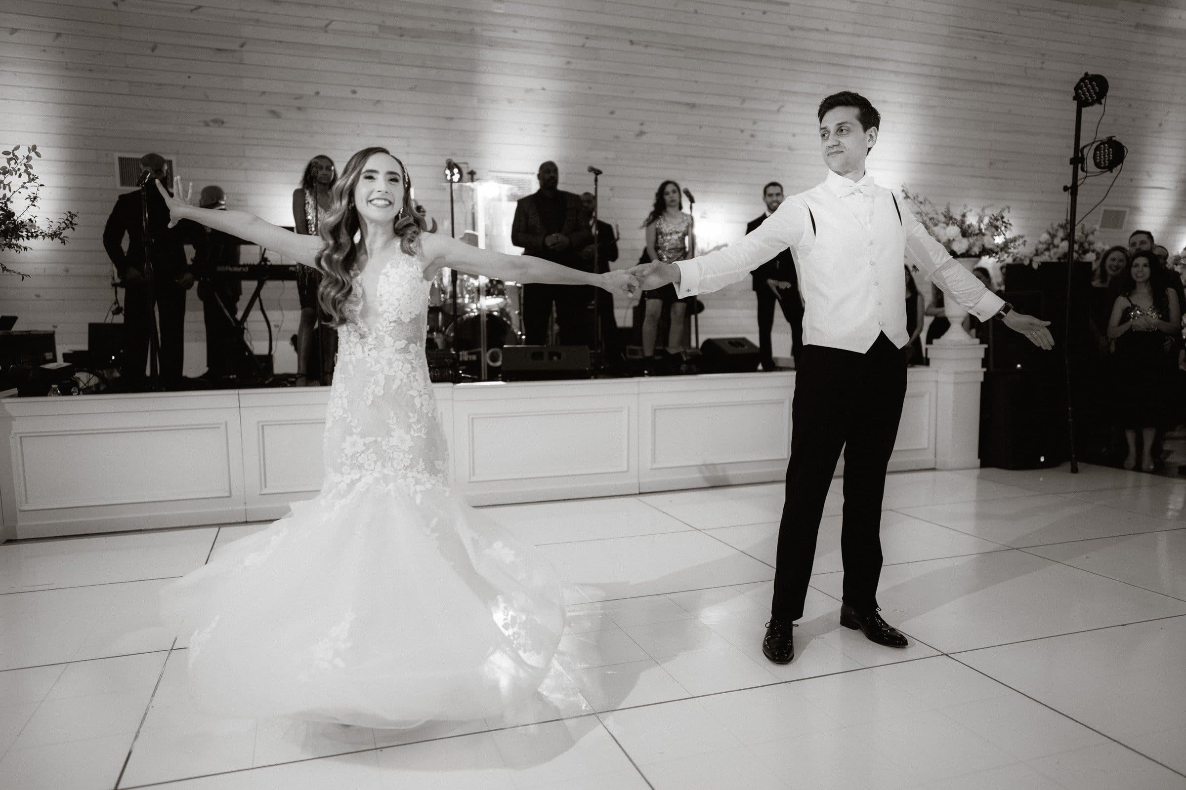 historic wakefield barn wedding photography - jennifer & stefan At a Wakefield barn wedding, a bride and groom joyfully dance on a white-tiled floor. She wears a flowing lace gown, and he is in a formal suit. A band and guests are visible in the softly lit background, captured beautifully by expert photography.