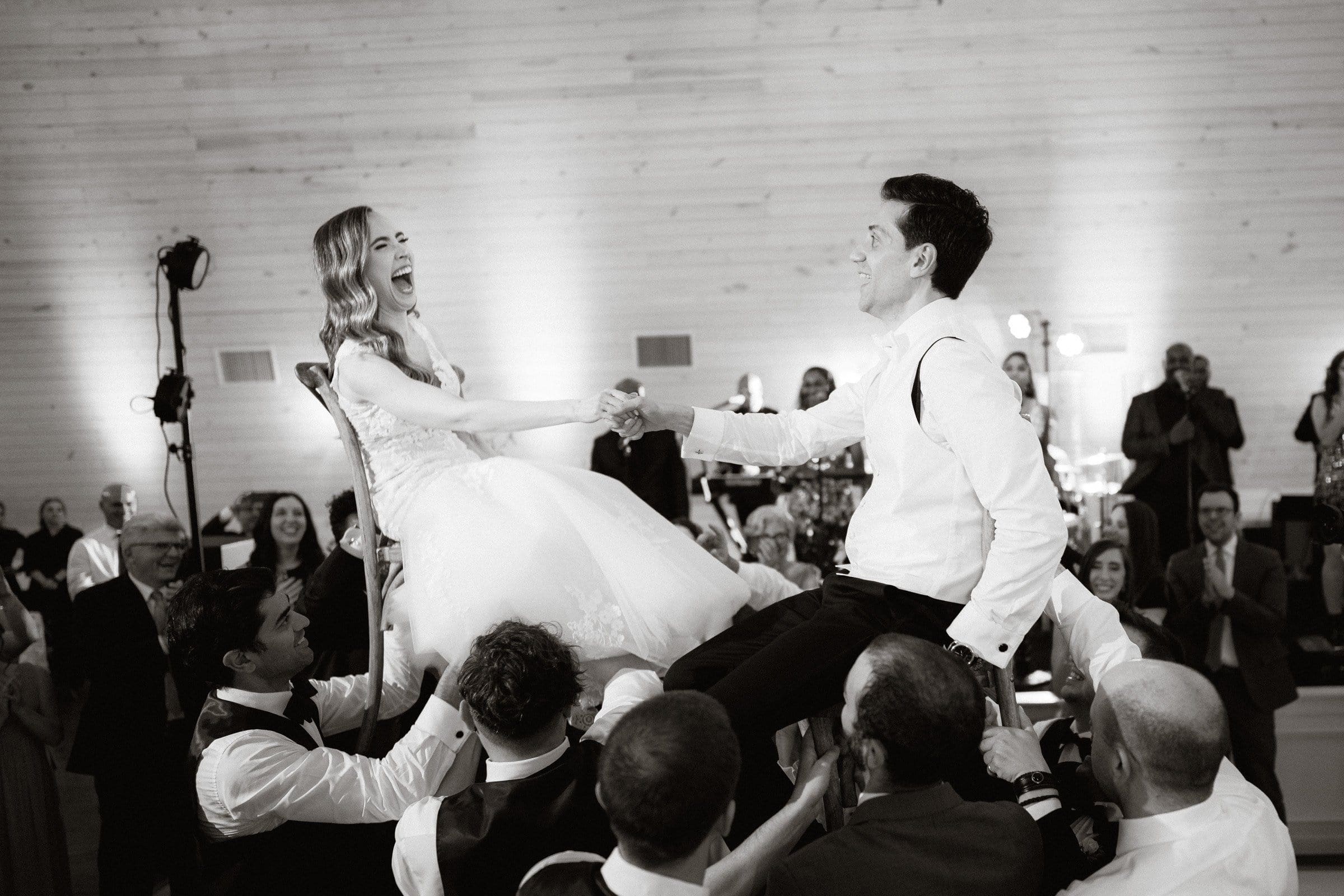 historic wakefield barn wedding photography - jennifer & stefan In the warmly lit venue of Wakefield Barn, a bride and groom are joyfully lifted on chairs by guests during a lively wedding celebration. With laughter and hands clasped tightly, they are surrounded by smiling attendees, capturing a moment of happiness and festivity.