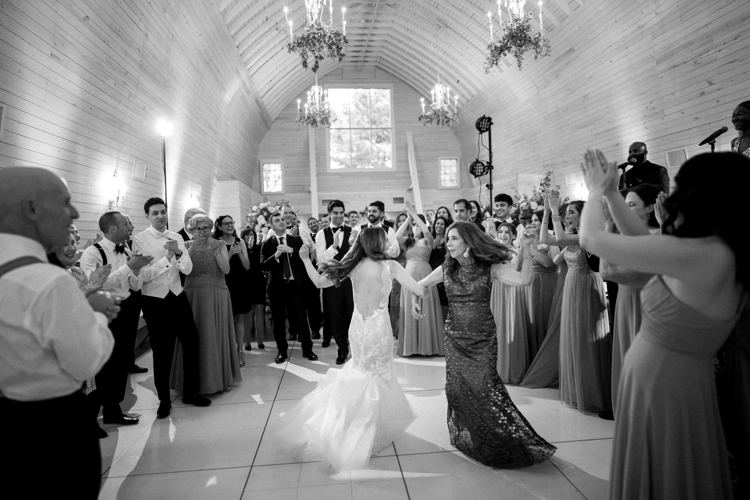 historic wakefield barn wedding photography - jennifer & stefan A bride and another woman dance joyfully in the center of a crowd at a wedding reception inside a wooden hall, as captured by Wakefield barn wedding photography. Guests in formal attire cheer and clap, creating a lively and celebratory atmosphere.