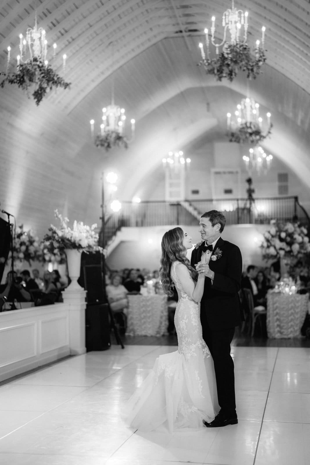 historic wakefield barn wedding photography - jennifer & stefan A couple dances elegantly in a grand ballroom adorned with chandeliers and floral arrangements. The bride, in a lace gown, and the groom, in a suit, create a picturesque scene worthy of Wakefield barn wedding photography, as guests watch their first dance from nearby tables.