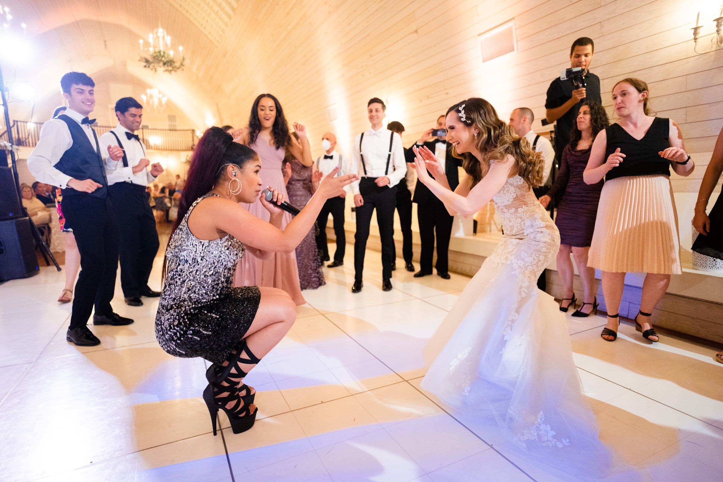 historic wakefield barn wedding photography - jennifer & stefan A bride in a white dress and a woman in sequined attire face each other on the dance floor, energetically squatting and gesturing while Wakefield Barn wedding photography captures the moment. A group of people surrounds them, some watching and others dancing, in a warmly lit hall.
