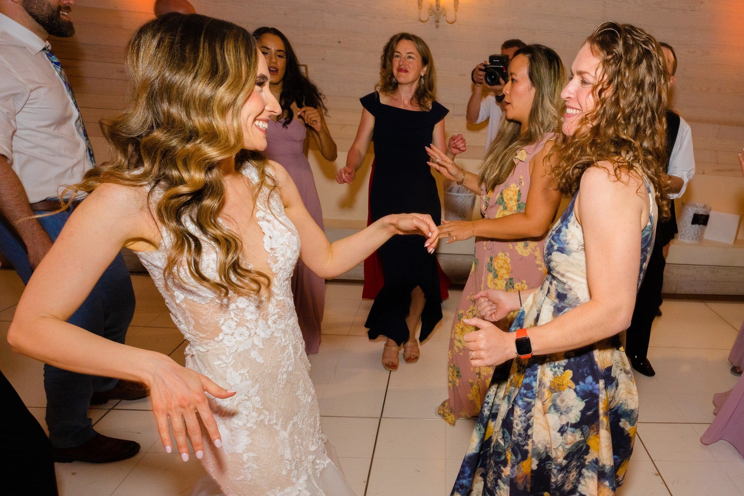 historic wakefield barn wedding photography - jennifer & stefan At a Wakefield barn wedding, a bride in a lace dress joyfully dances with a guest in floral attire. Other guests are dancing and mingling in the background, capturing the lively celebration atmosphere beautifully through stunning photography.
