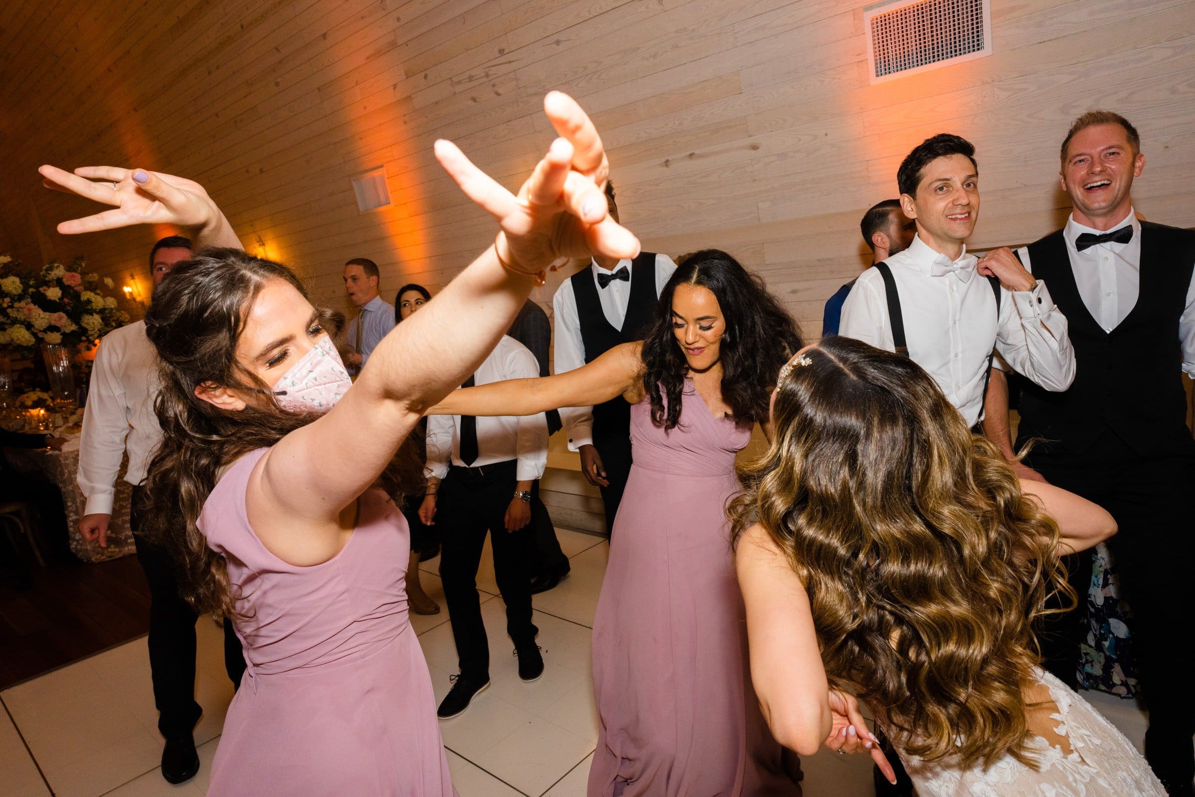 historic wakefield barn wedding photography - jennifer & stefan At a Wakefield barn wedding, guests joyfully dance and celebrate. Two women in lavender dresses and a woman in white laugh and raise their arms, as a group of men in white shirts and bow ties join the fun. The mood is lively and festive, perfect for capturing unforgettable wedding photography moments.