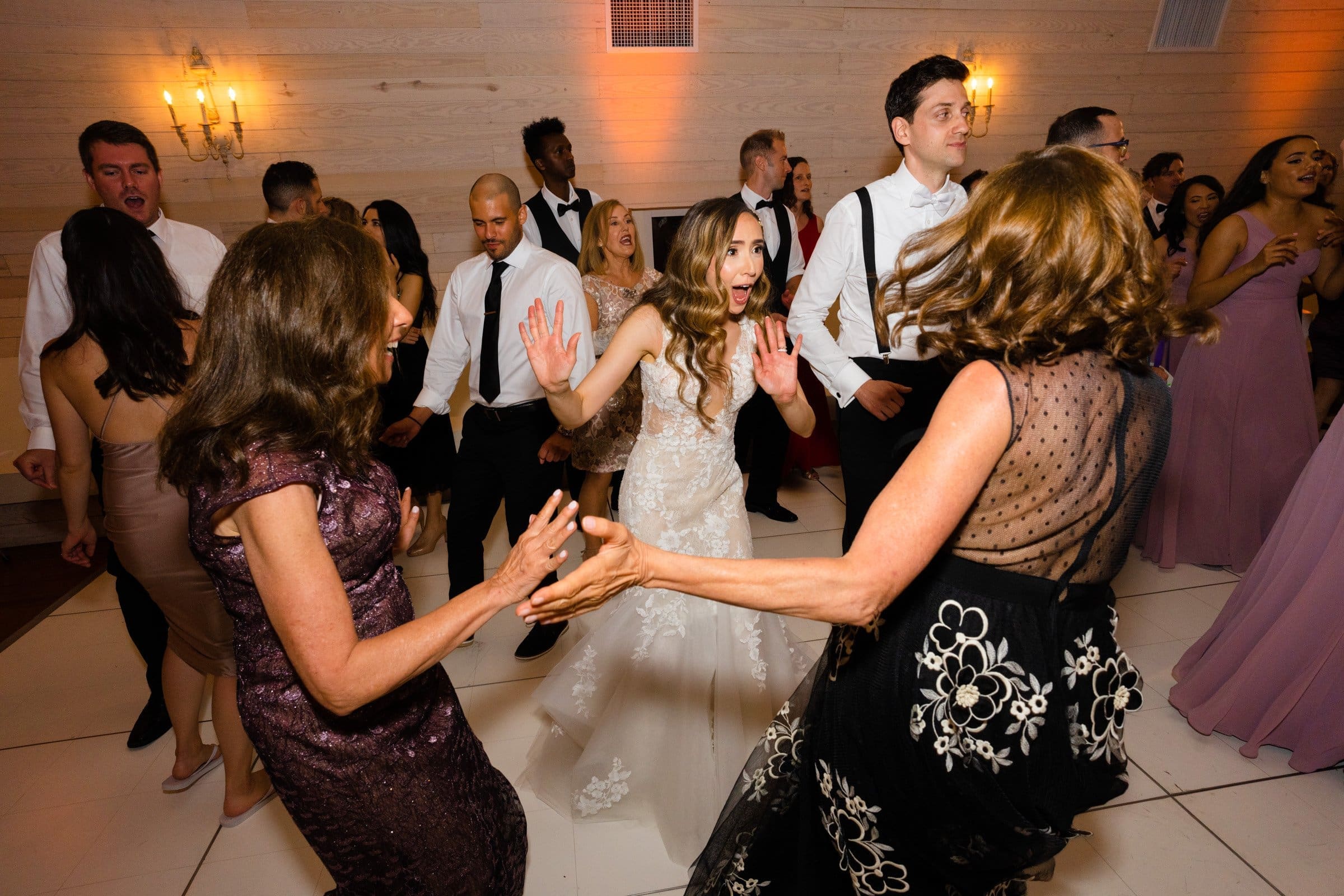 historic wakefield barn wedding photography - jennifer & stefan A bride in a white gown dances energetically with two women in elegant dresses. Surrounded by guests, some clapping and others dancing, at the warmly lit reception hall of Wakefield Barn, the atmosphere captured through wedding photography is joyful and lively.