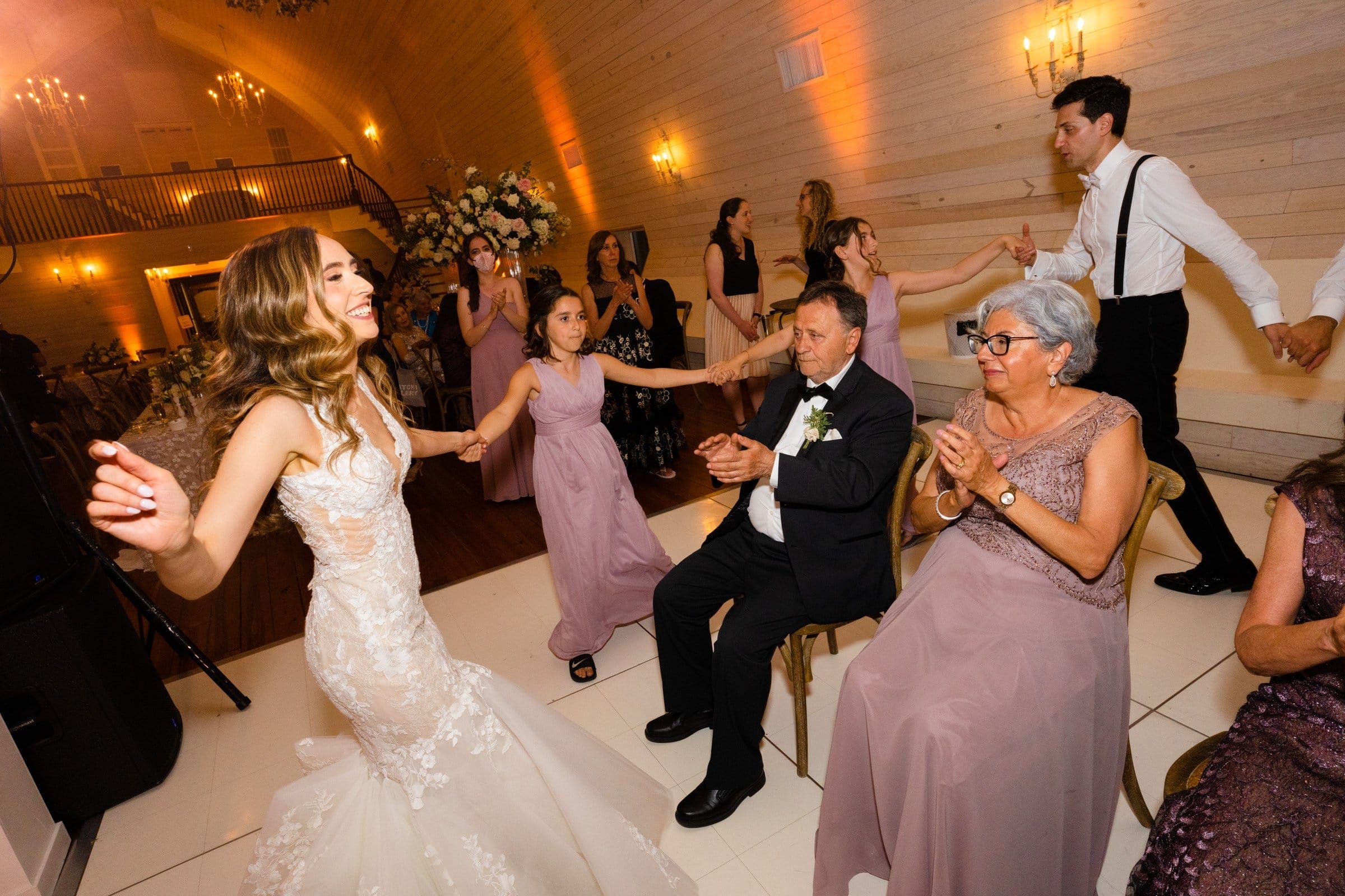 historic wakefield barn wedding photography - jennifer & stefan Bride dancing joyfully with guests at a Wakefield barn wedding reception. She's holding hands and smiling. Several guests, including a seated elderly woman and a man in formal wear, clap and enjoy the celebration. The warmly lit room is elegantly decorated, perfect for stunning photography.