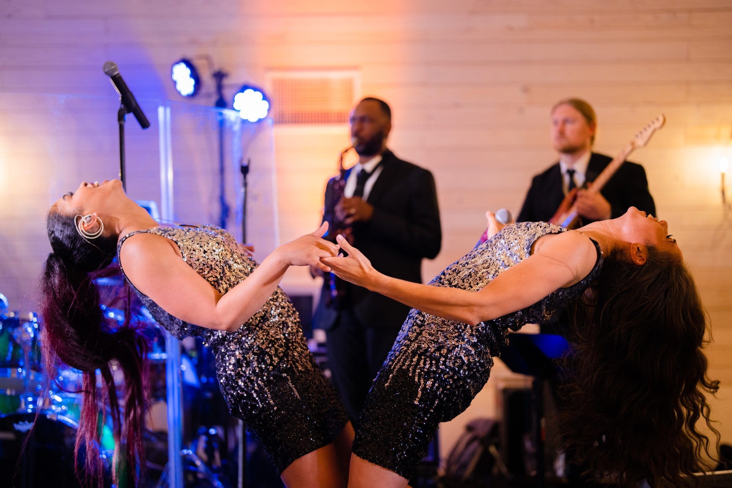 historic wakefield barn wedding photography - jennifer & stefan Two performers in sequined outfits lean back dramatically while singing on stage, capturing the essence of a Wakefield barn wedding. A band with a guitarist and saxophonist plays in the background, set against a rustic wooden wall enhanced by vibrant stage lighting.