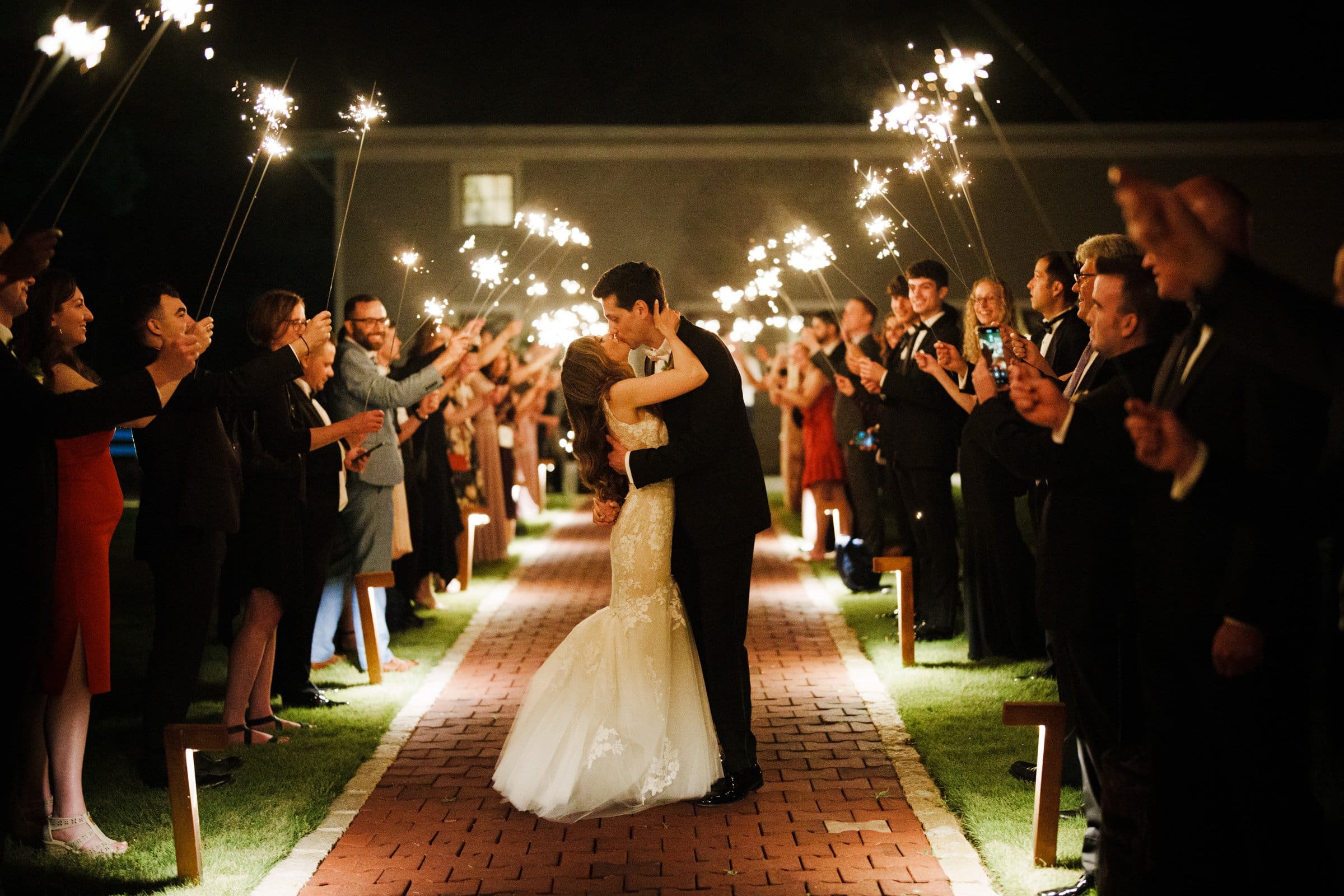 historic wakefield barn wedding photography - jennifer & stefan At a Wakefield barn wedding, a newlywed couple shares a kiss on the brick path at night, surrounded by guests holding sparklers. The guests line each side, crafting a celebratory and festive atmosphere under the shimmering night sky, captured in stunning photography.