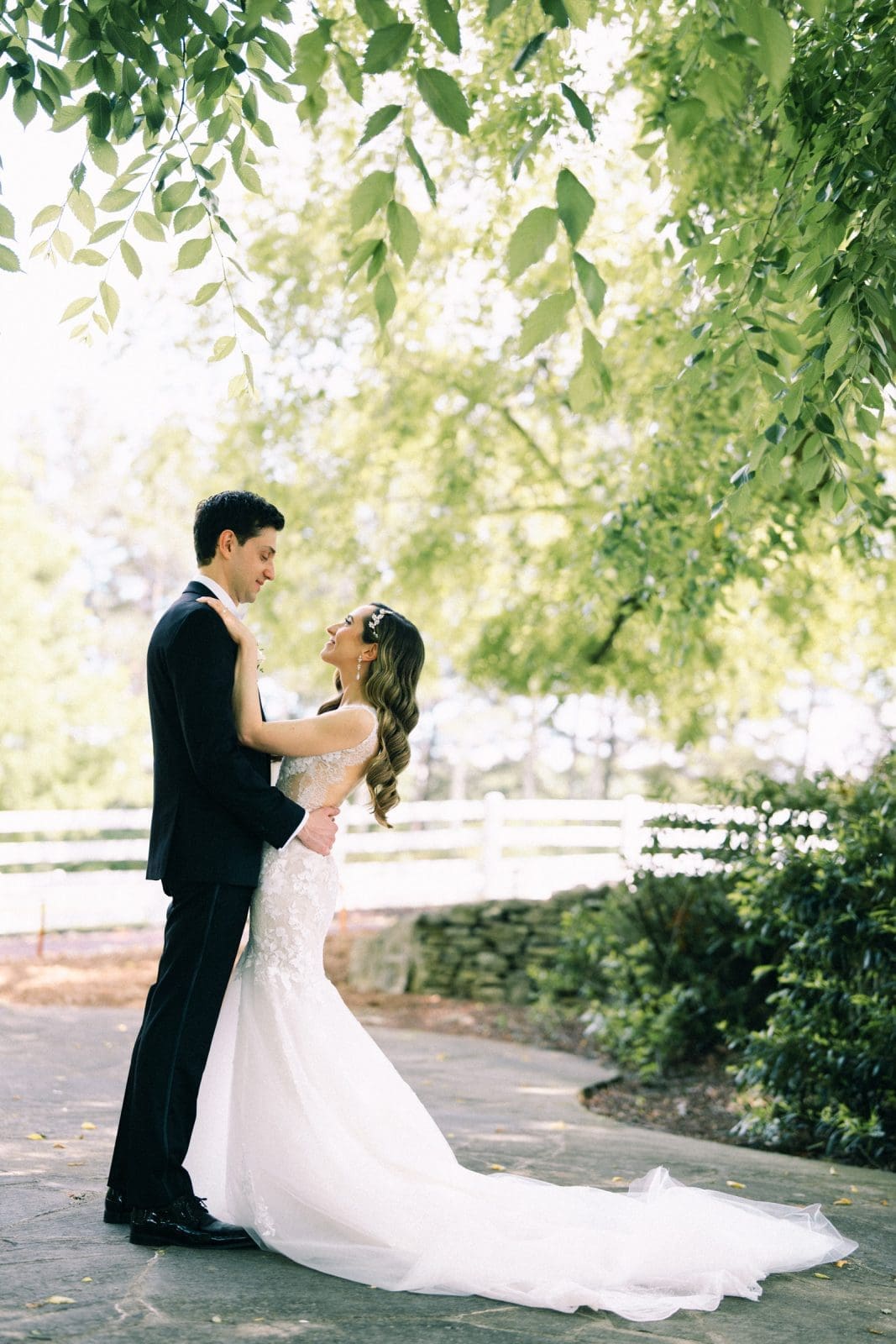 historic wakefield barn wedding photography - jennifer & stefan A bride and groom stand facing each other under a tree at Wakefield Barn, surrounded by lush greenery. The bride wears a white dress and the groom a dark suit. They gaze at each other lovingly, with sunlight filtering through the leaves—a perfect moment captured in stunning wedding photography.