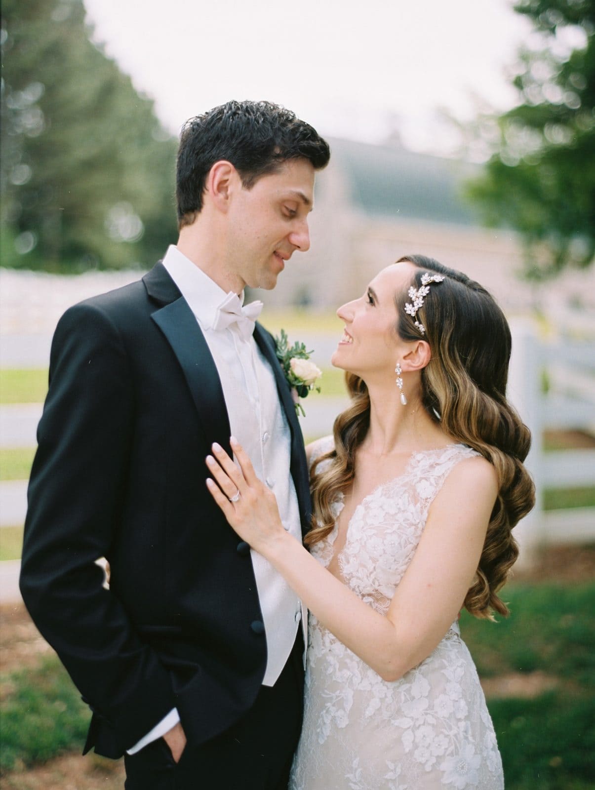 historic wakefield barn wedding photography - jennifer & stefan A bride and groom lovingly gaze at each other outdoors, captured in stunning Wakefield barn wedding photography. The bride wears an elegant lace gown with a jeweled hair accessory, while the groom dons a black tuxedo and white bow tie. They stand before a white fence, greenery providing the perfect backdrop.