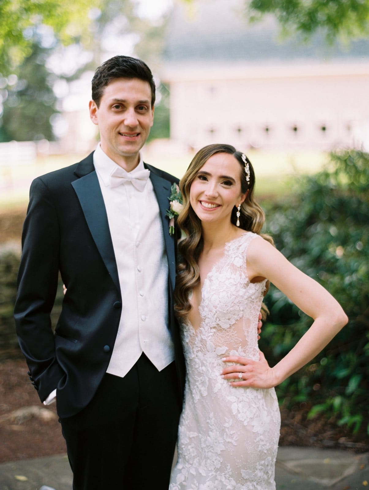historic wakefield barn wedding photography - jennifer & stefan In the lush garden of Wakefield Barn, a bride in a lace dress wraps her arm lovingly around the groom, who stands in a dashing tuxedo with a white bow tie and boutonniere. Their smiles radiate joy, set against a backdrop of blurred greenery captured in exquisite wedding photography.