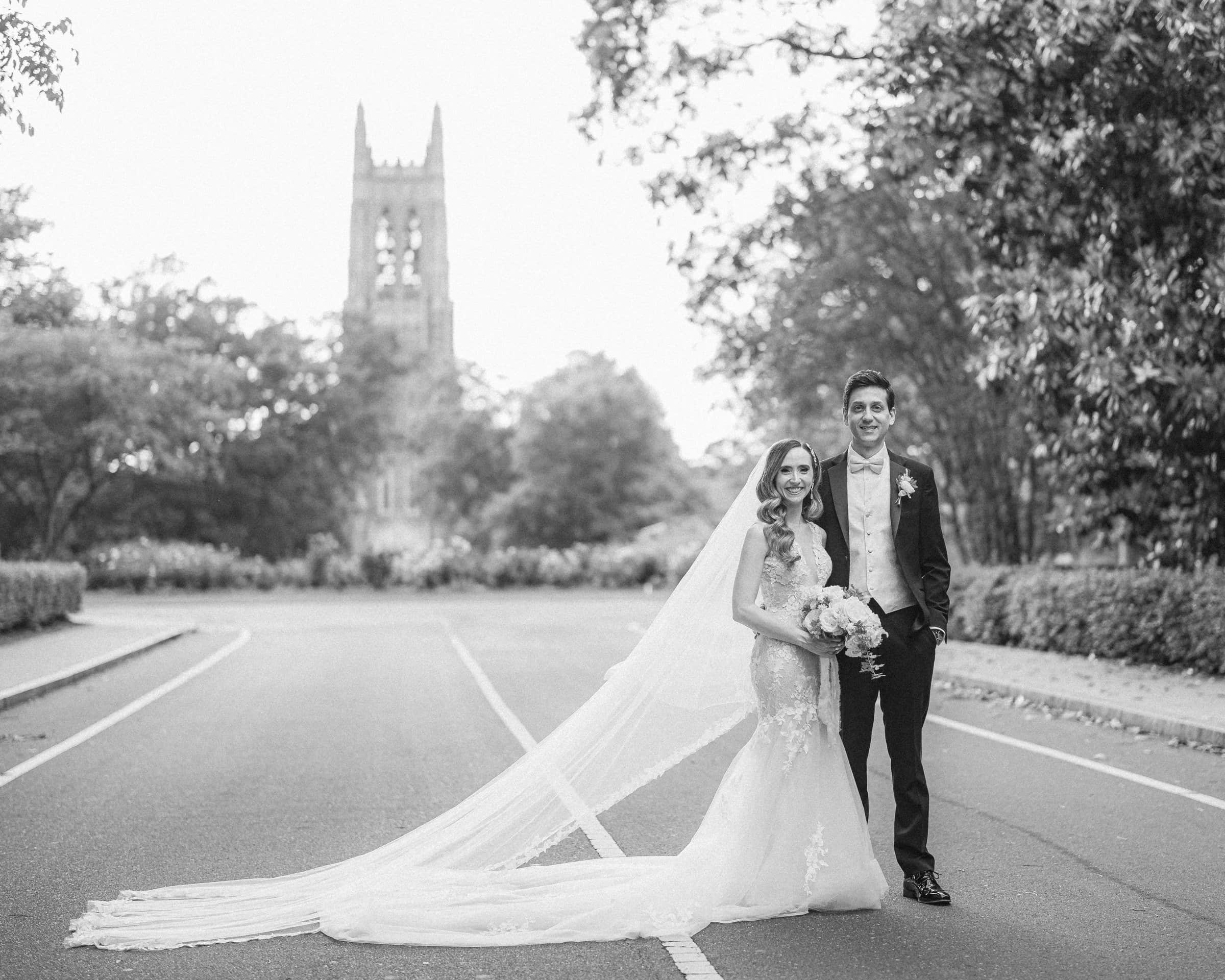 historic wakefield barn wedding photography - jennifer & stefan A bride and groom stand on a road, the groom's hand around the bride's waist, as her veil trails behind her. Trees line the road, and a tall tower looms in the background. Captured in black and white, this imagery embodies timeless Wakefield barn wedding photography.