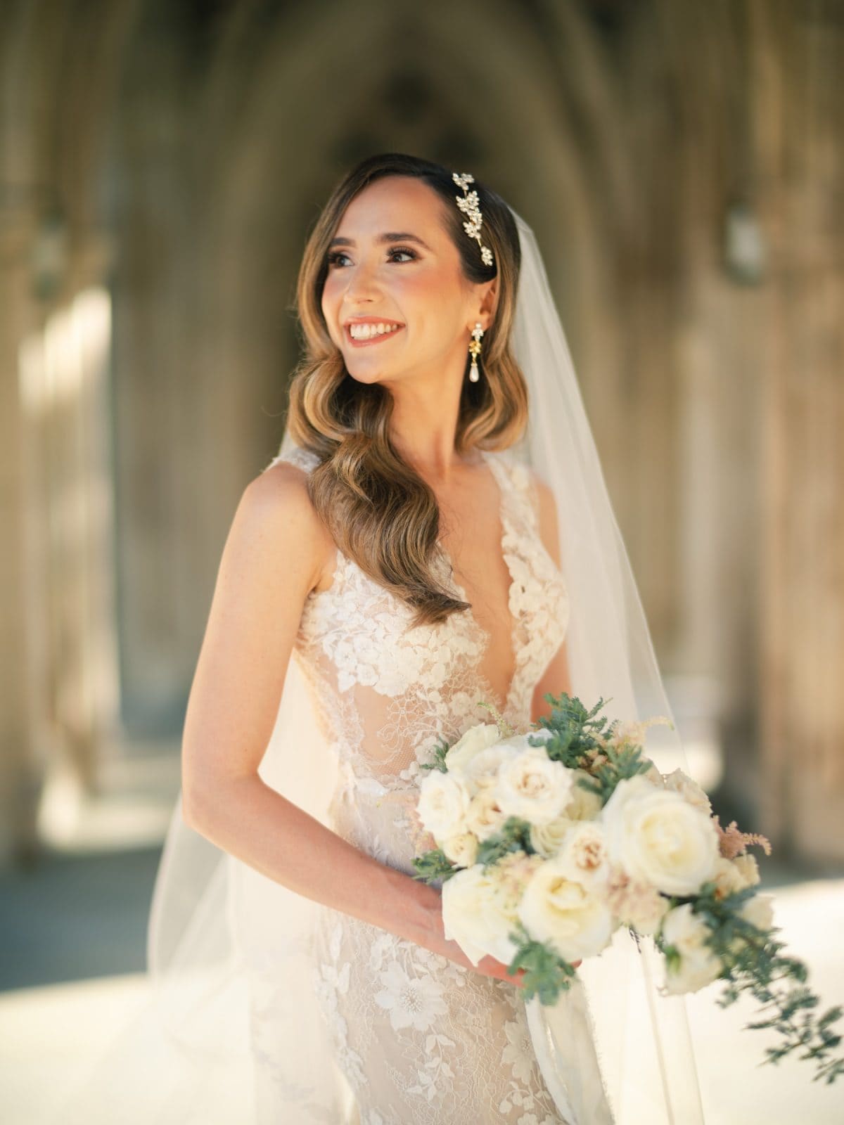 historic wakefield barn wedding photography - jennifer & stefan A bride in a lace wedding dress smiles while holding a bouquet of white and pastel flowers at a picturesque Wakefield barn wedding. Her long wavy hair, adorned with a decorative hairpin, complements her veil. The blurred arches in the background create a soft, elegant setting perfect for photography.