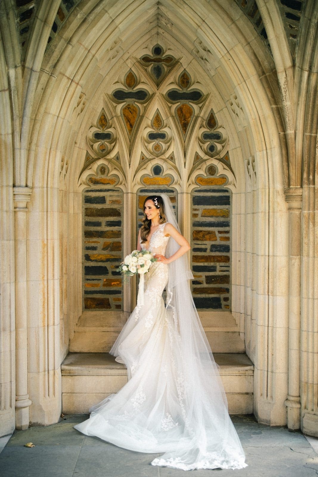 historic wakefield barn wedding photography - jennifer & stefan A bride in a long, elegant lace wedding gown and veil stands in a stone archway at Wakefield Barn. She holds a bouquet of white roses and looks to the side, surrounded by ornate Gothic architecture that adds a timeless charm to the wedding photography setting.