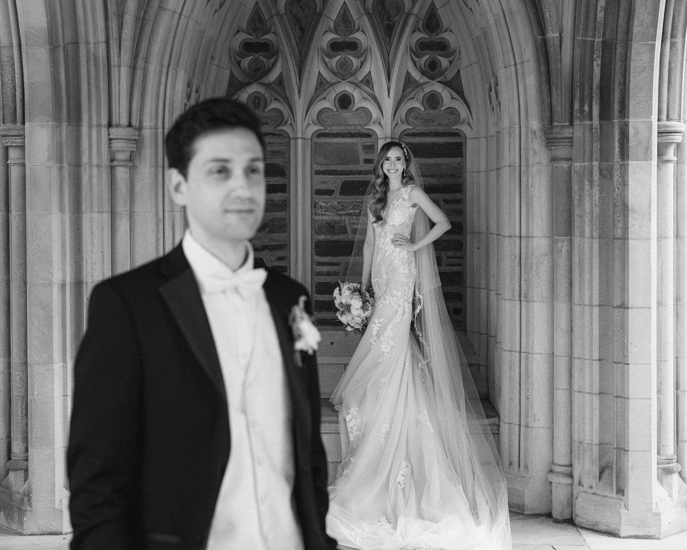 historic wakefield barn wedding photography - jennifer & stefan A bride and groom are pictured at their Wakefield barn wedding. The groom, in focus, stands in the foreground wearing a tuxedo, while the bride, slightly out of focus, smiles and holds a bouquet in front of an ornate stone archway.