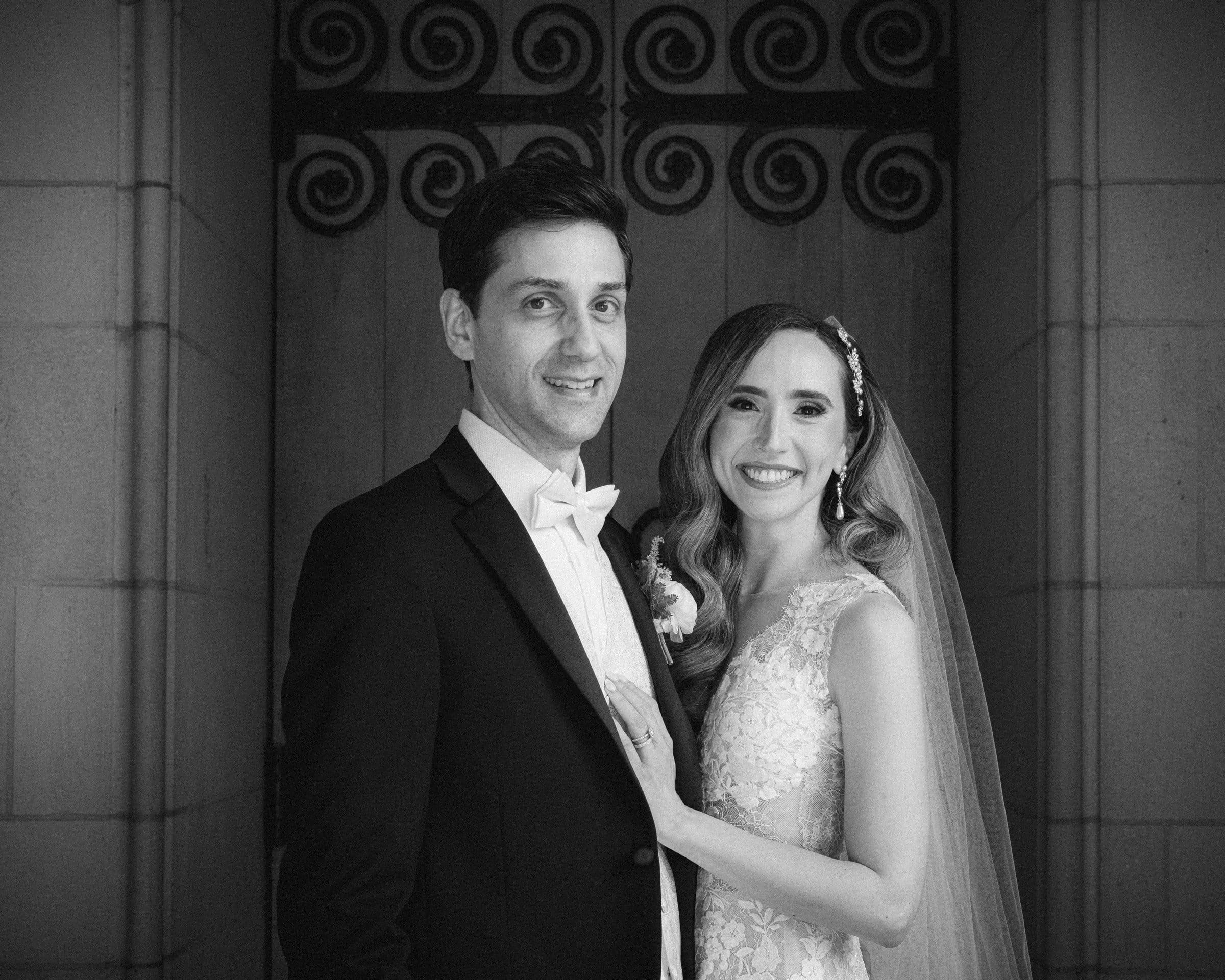 historic wakefield barn wedding photography - jennifer & stefan A black and white photo captures a bride and groom at their Wakefield barn wedding. The groom dons a suit with a bow tie, while the bride, in her lace dress and veil, smiles. They stand intimately together before a decorative iron gate.