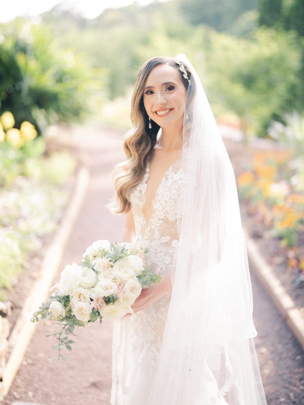 historic wakefield barn wedding photography - jennifer & stefan A bride in a white lace dress and veil smiles while holding a bouquet of white and pale pink flowers. She stands on a garden path surrounded by greenery under soft natural light, captured beautifully in Wakefield barn wedding photography.