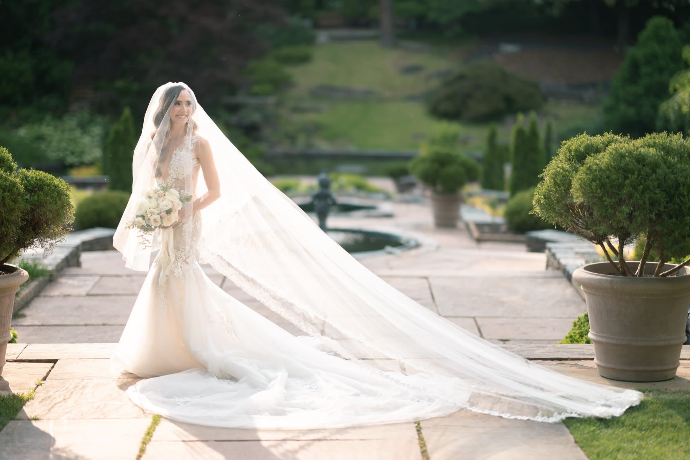 historic wakefield barn wedding photography - jennifer & stefan A bride in a long, flowing white gown and veil holds a bouquet of white flowers, captured beautifully in Wakefield barn wedding photography. She stands outdoors on a stone path surrounded by lush greenery and neatly trimmed potted plants, with a blurred garden landscape in the background.