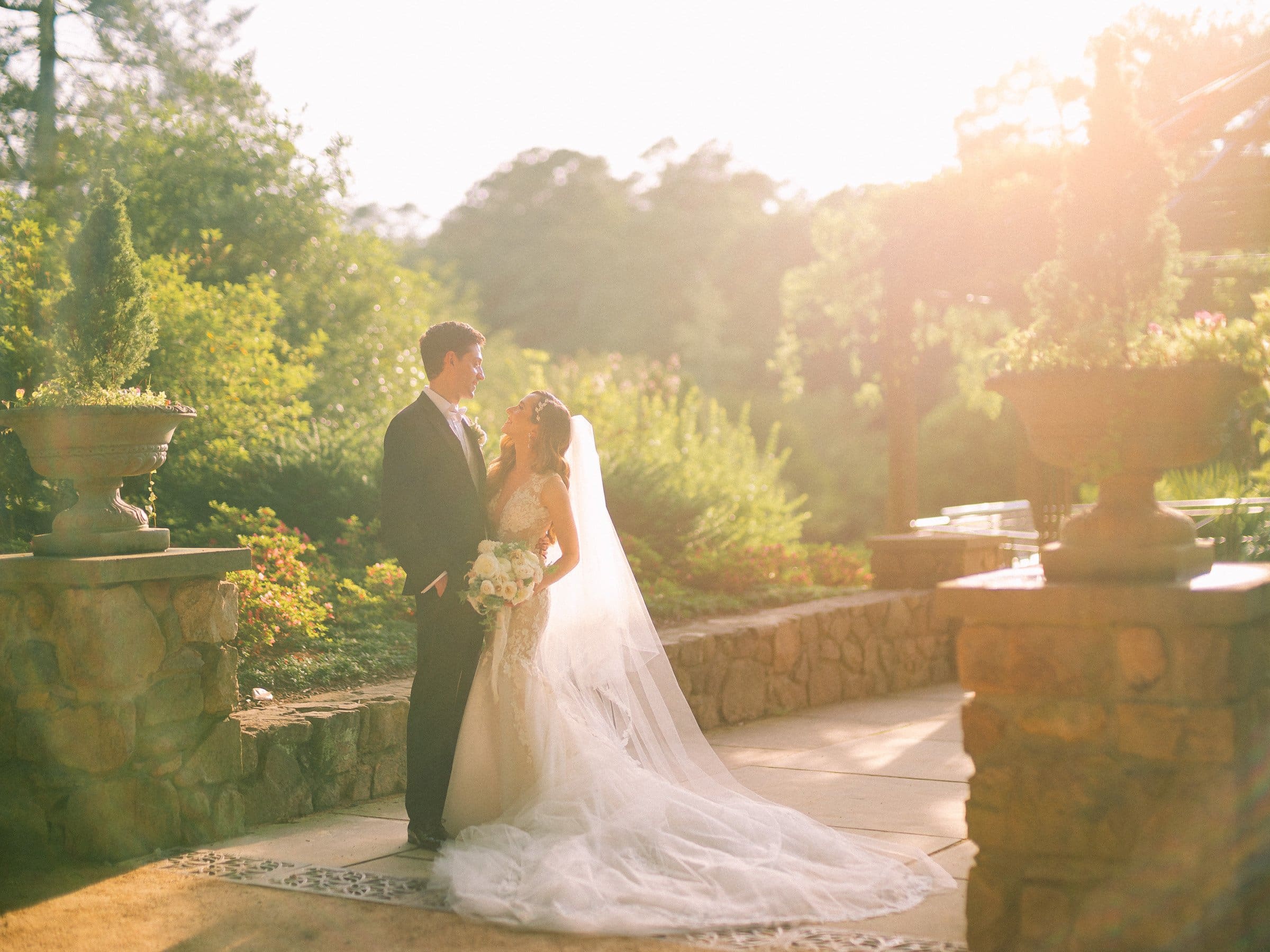 historic wakefield barn wedding photography - jennifer & stefan In the sunlit garden of Wakefield, a bride and groom stand surrounded by stone walls and lush greenery. The bride, in a long white gown with a flowing veil, holds a bouquet as they gaze at each other lovingly, capturing the essence of barn wedding photography.