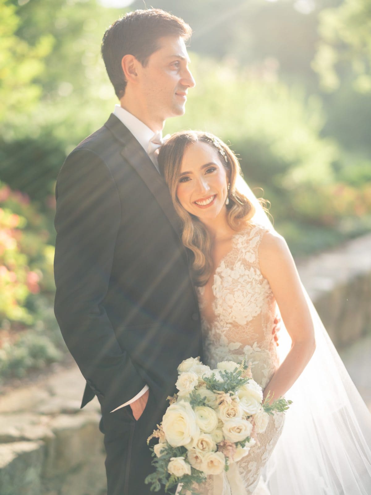 historic wakefield barn wedding photography - jennifer & stefan A bride and groom stand in a sunlit garden at Wakefield Barn. The bride wears a lace wedding dress and holds a bouquet of white and blush roses, while the groom sports a black suit. Both are smiling, bathed in soft, natural light, capturing the essence of wedding photography.