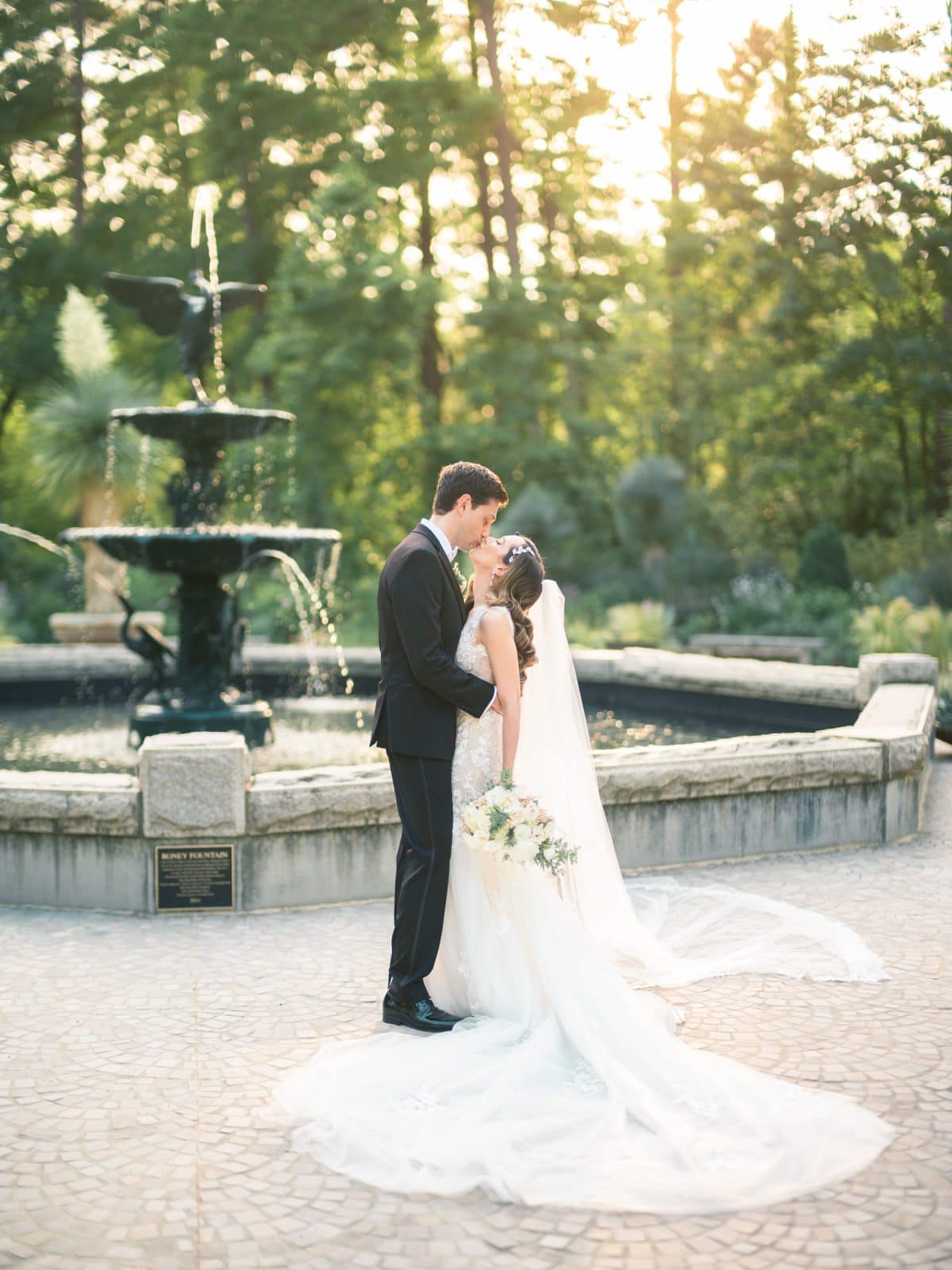 historic wakefield barn wedding photography - jennifer & stefan A bride and groom share a kiss in front of a large, elegant fountain at their Wakefield barn wedding. The bride holds a bouquet and wears a long gown with a train, while the groom is in a dark suit. The sun sets in the background, casting a warm glow over the enchanting scene.