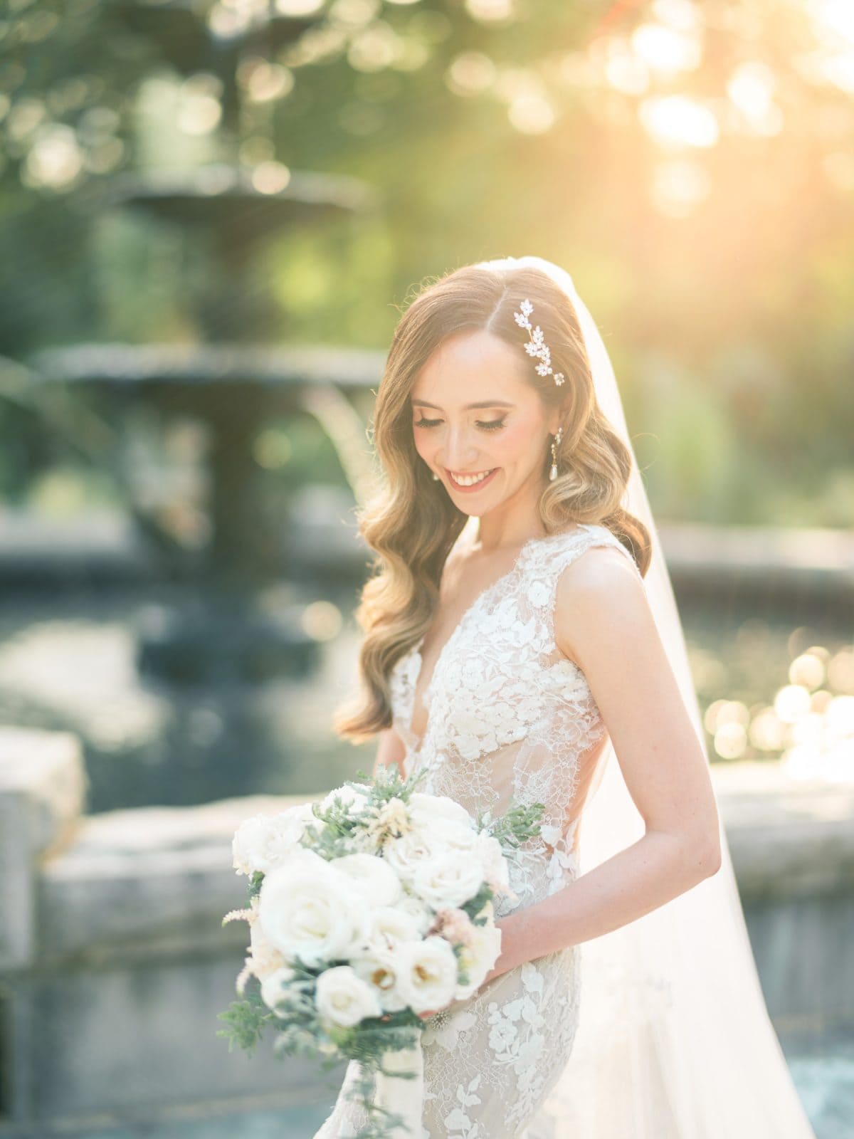 historic wakefield barn wedding photography - jennifer & stefan A smiling bride in a lace wedding dress stands by a fountain at Wakefield Barn. She holds a bouquet of white and blush flowers as the sun shines softly, creating a warm, ethereal glow perfect for wedding photography.