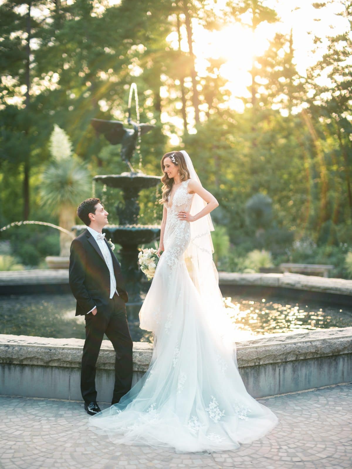 historic wakefield barn wedding photography - jennifer & stefan A bride and groom pose by a fountain in a sunlit garden at Wakefield Barn. The bride stands on the fountain edge in a flowing white dress, holding a bouquet. The groom, in a black suit, stands nearby, looking at her. Sunlight filters through the trees in the background, capturing perfect wedding photography moments.