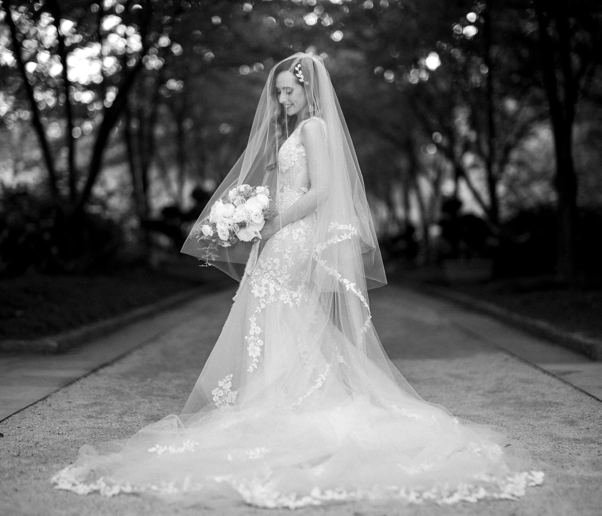 A bride stands on a path in a forested area, wearing a lace wedding dress and veil, and holding a bouquet. The black-and-white photo, reminiscent of Wakefield barn wedding photography, captures her in a serene, natural setting.