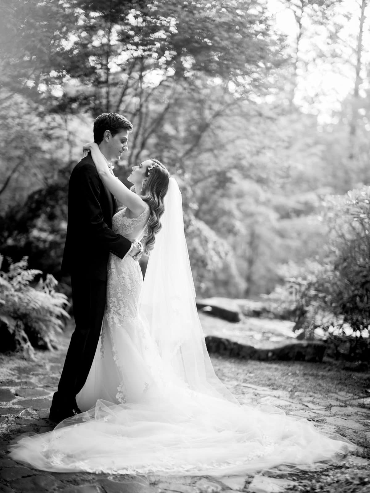 historic wakefield barn wedding photography - jennifer & stefan In a serene, forested setting reminiscent of Wakefield barn wedding photography, a bride in a flowing gown with a long veil embraces her groom in a dark suit. Sunlight filters through the trees, creating a dreamy, romantic atmosphere.