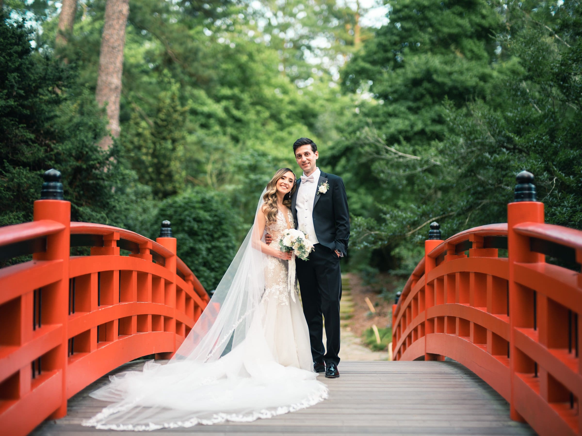 historic wakefield barn wedding photography - jennifer & stefan A bride and groom stand together on a red wooden bridge surrounded by lush greenery at Wakefield Barn. The bride holds a bouquet and wears a long, flowing veil, while the groom is in a black suit. They both look content and happy, captured perfectly in this wedding photography moment.