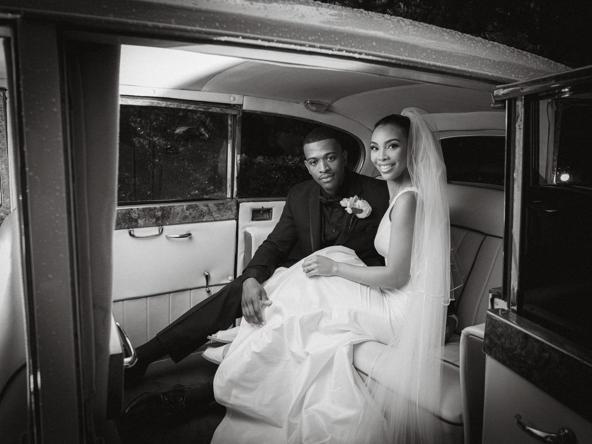 umstead wedding photography - kayla & deandre A bride and groom sit inside a classic car at their Umstead wedding. The bride, in her white dress and veil, smiles as the groom beside her, in a dark suit, looks at the camera. The elegant interior enhances the vintage, romantic mood of this special day.