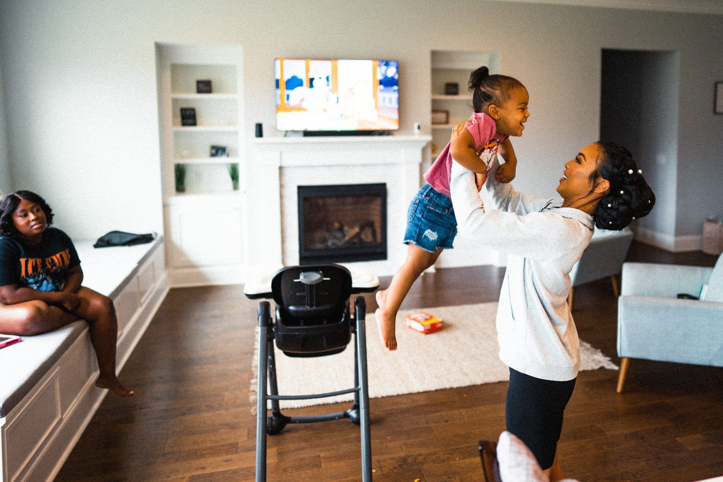 umstead wedding photography - kayla & deandre A woman playfully lifts a smiling child in a cozy living room, reminiscent of an Umstead wedding's joyful atmosphere. Another person sits on a bench nearby. The room features a TV above the fireplace, with toy blocks and a highchair scattered on the floor.