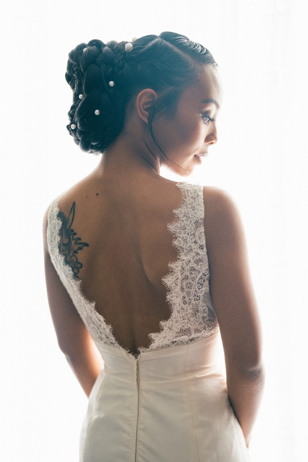 umstead wedding photography - kayla & deandre A woman in a white lace wedding dress gazes over her shoulder at an enchanting Umstead wedding. Her hair is styled elegantly with pearl accents, and a small tattoo graces her upper back. The background is softly lit, capturing the romance of the moment.