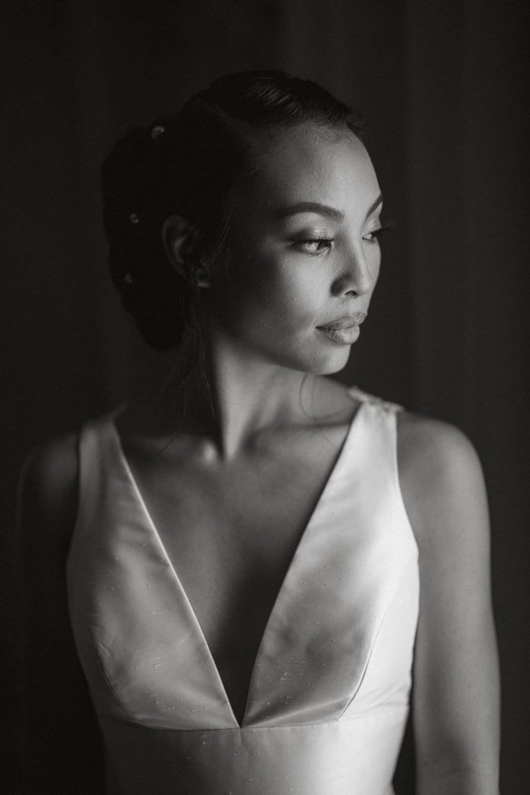 umstead wedding photography - kayla & deandre A bride in a white dress looks to the side. The black and white image emphasizes her serene expression and the elegant simplicity of her attire, perfect for an Umstead wedding. Her hair is styled in an updo, with lighting creating a soft, dramatic effect.