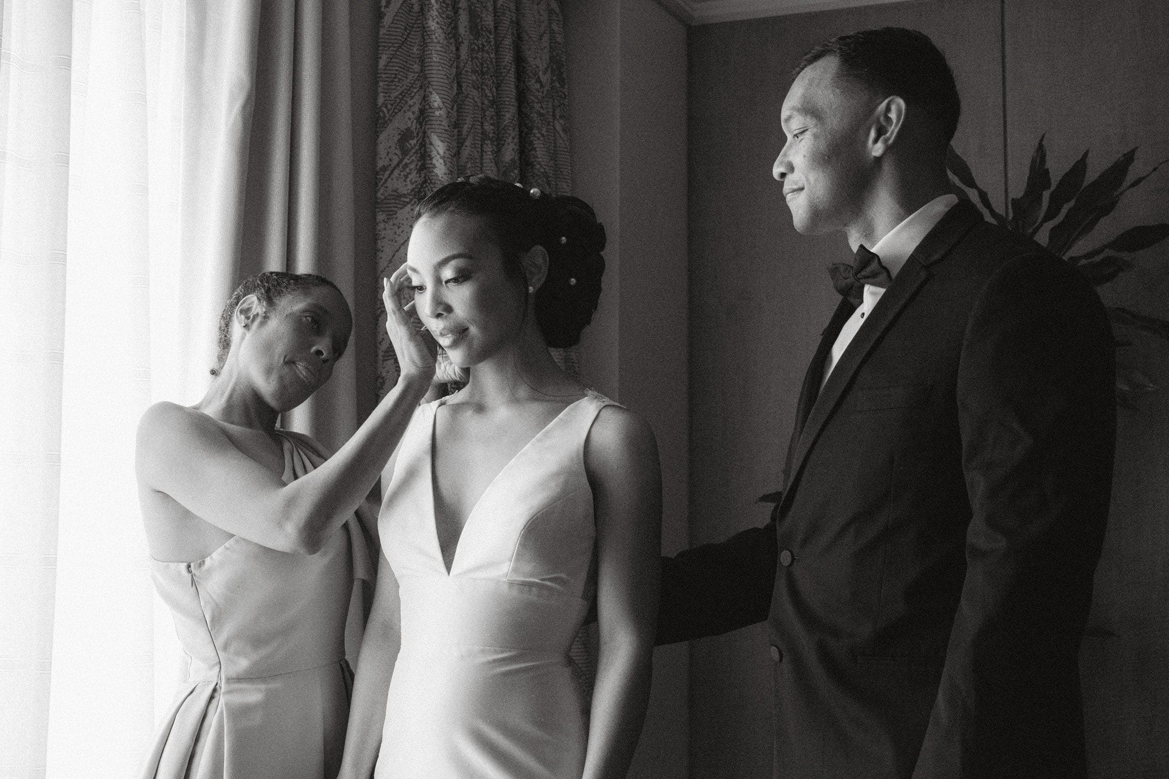 umstead wedding photography - kayla & deandre In an elegant Umstead wedding, a black and white photo captures a bride in her wedding dress standing beside two people. One person gently touches her face while the other, dressed in a suit, stands close and smiles at her. The backdrop features graceful curtains and lush plants.