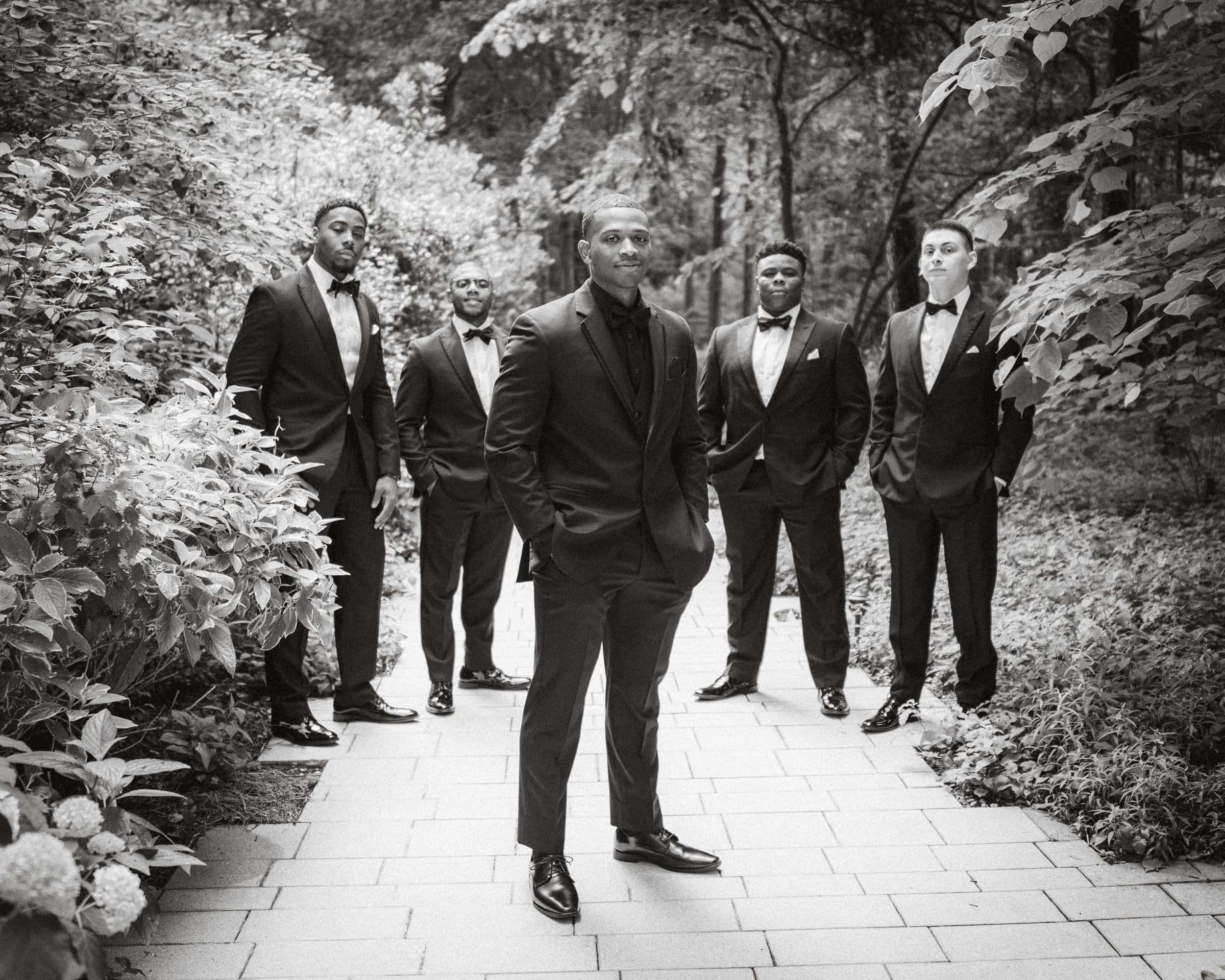 umstead wedding photography - kayla & deandre At an Umstead wedding, a group of five men in formal black suits and bow ties stand on a stone path in a garden. Four men are slightly behind the central figure, who stands confidently with his hands in his pockets. Lush greenery surrounds them, captured beautifully in black and white.