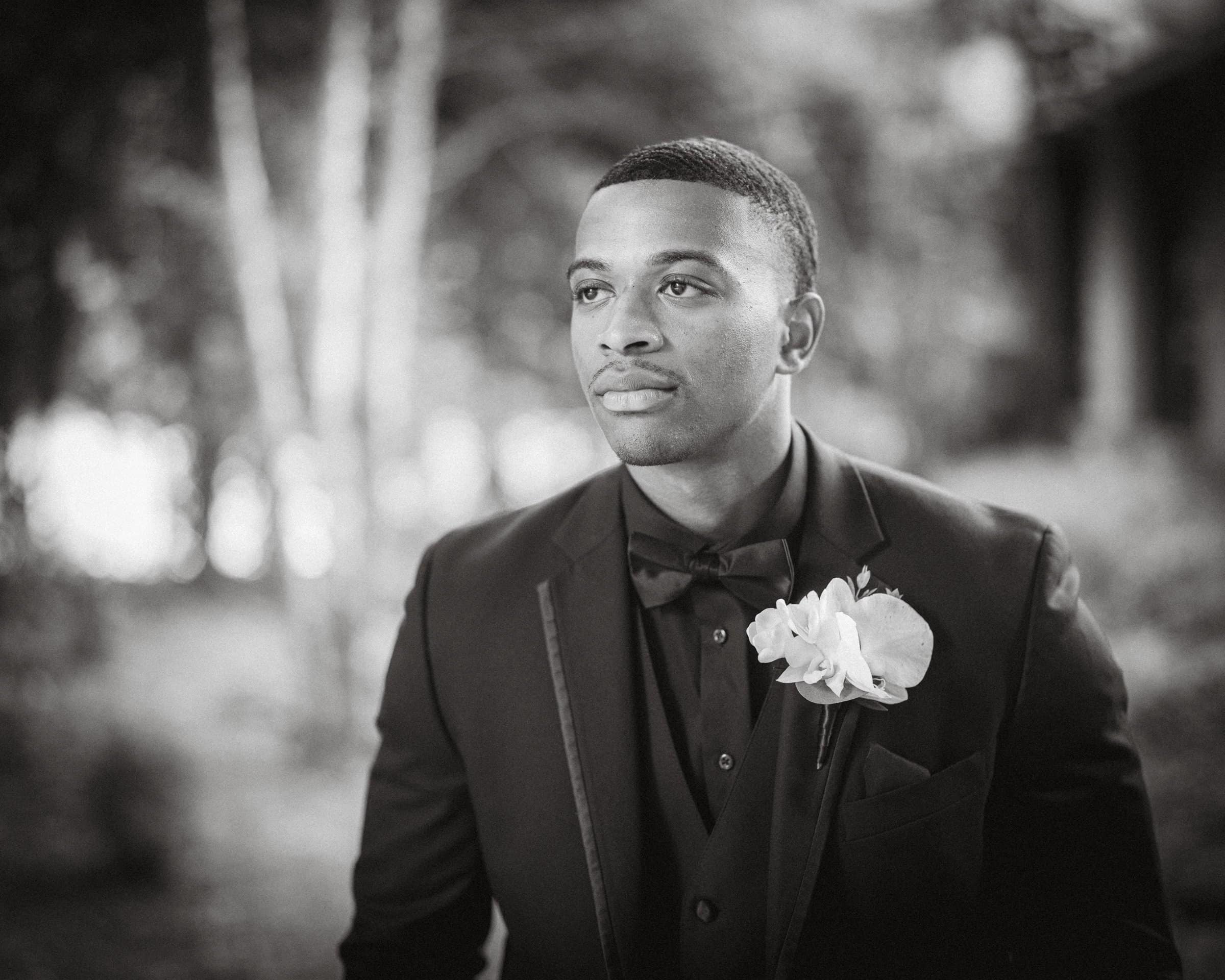 umstead wedding photography - kayla & deandre A man in a formal suit with a bow tie and a flower boutonniere gazes thoughtfully to the side, reminiscent of an Umstead wedding. The background is blurred with trees and foliage, creating a soft and contemplative atmosphere. The image is in black and white.