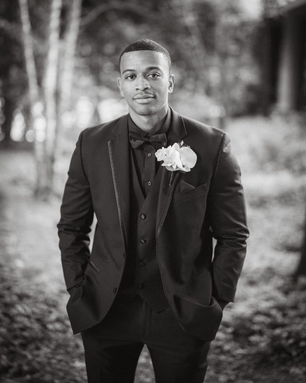 umstead wedding photography - kayla & deandre In this black and white photo, a man in a formal suit stands outdoors at an Umstead wedding, his hands in pockets. Wearing a bow tie and a white flower pinned to his lapel, the softly blurred trees and foliage form an elegant backdrop.