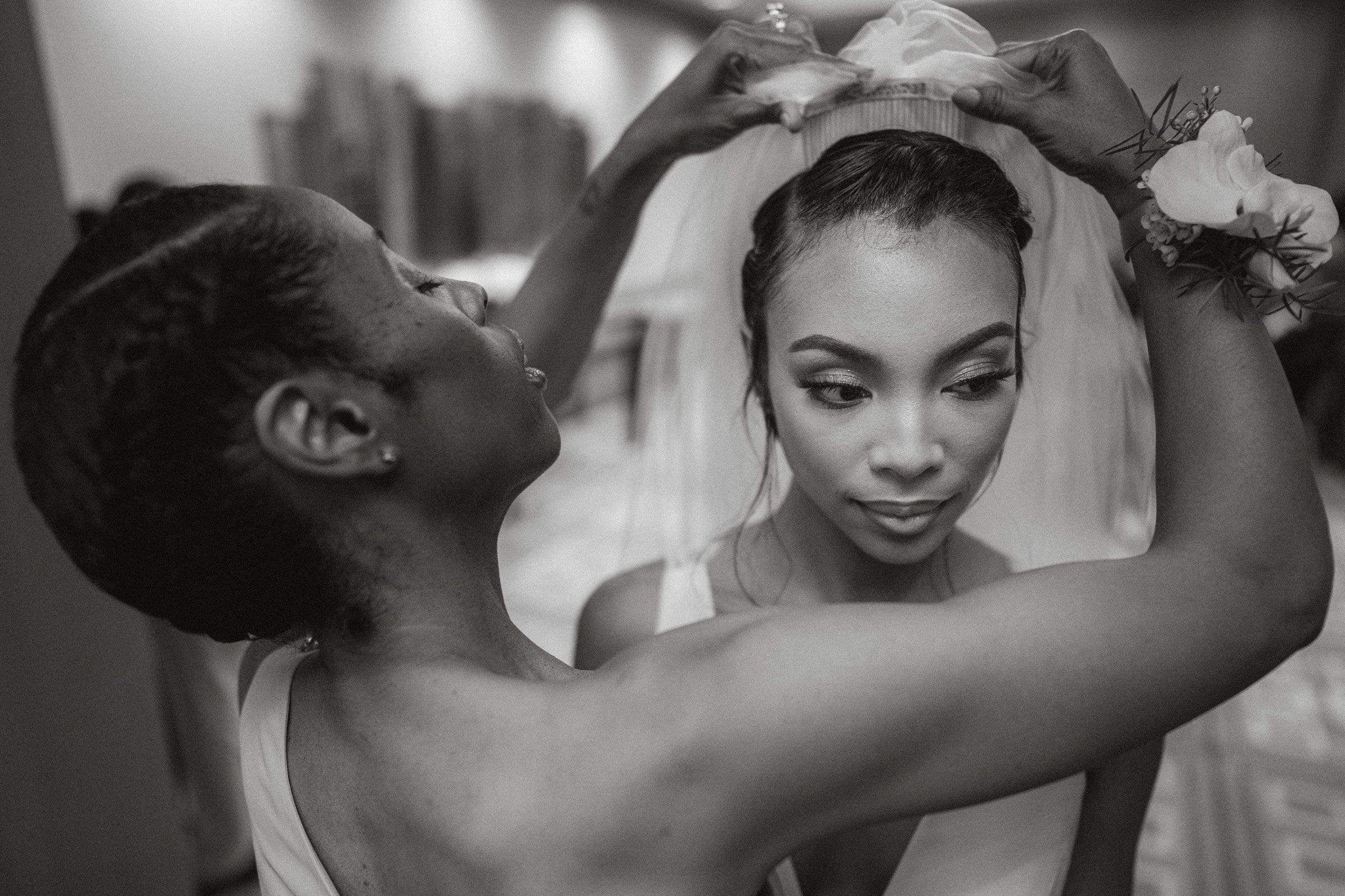umstead wedding photography - kayla & deandre In a timeless black and white photo from an Umstead wedding, a bride prepares with grace. Another woman delicately adjusts her veil, highlighting the intricate updo and elegant gown. Their focused expressions capture the essence of this cherished moment of preparation.
